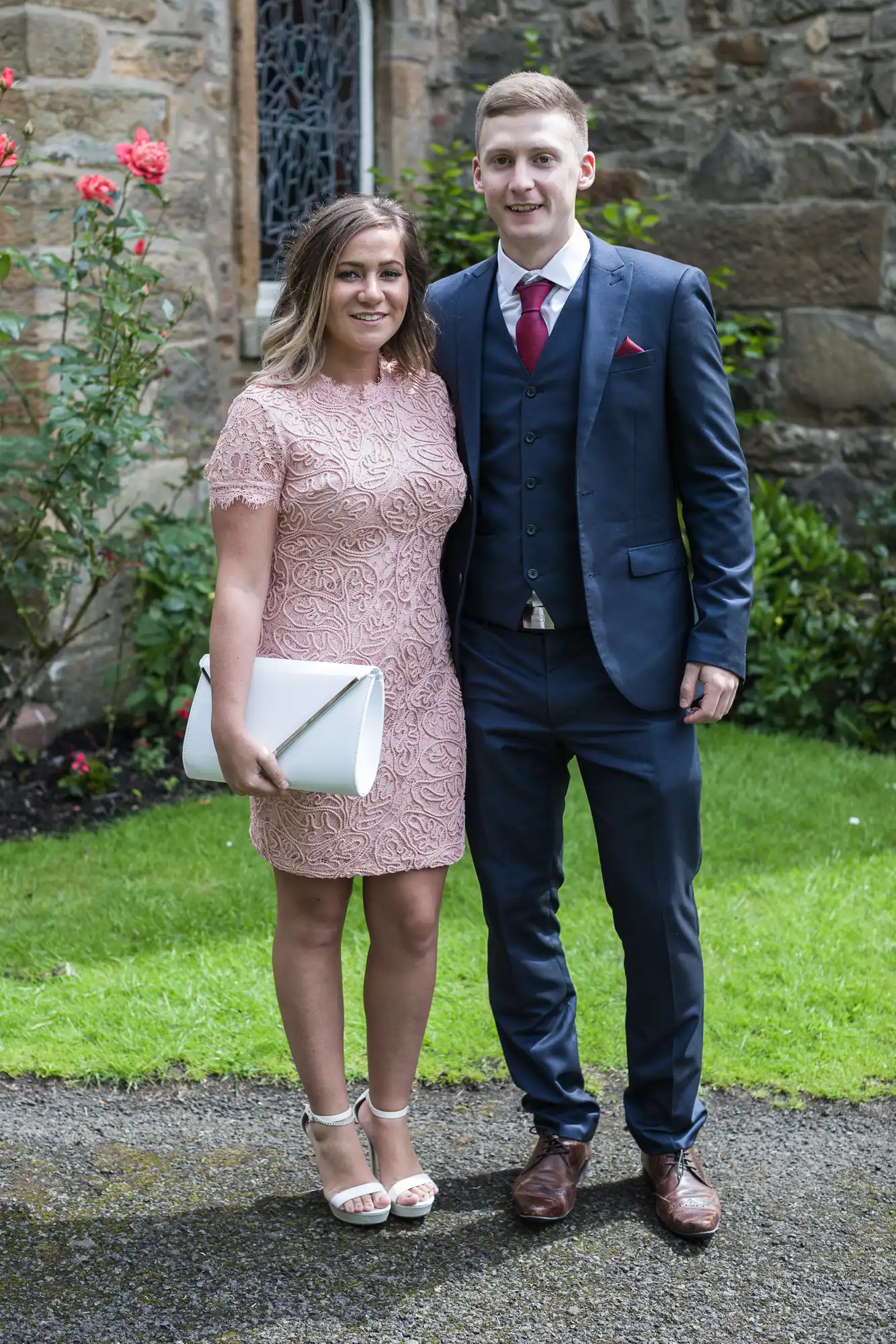 A woman in a pink lace dress and white heels stands next to a man in a dark blue suit and red tie. They are outdoors on a grassy area with a stone building and rose bushes in the background.