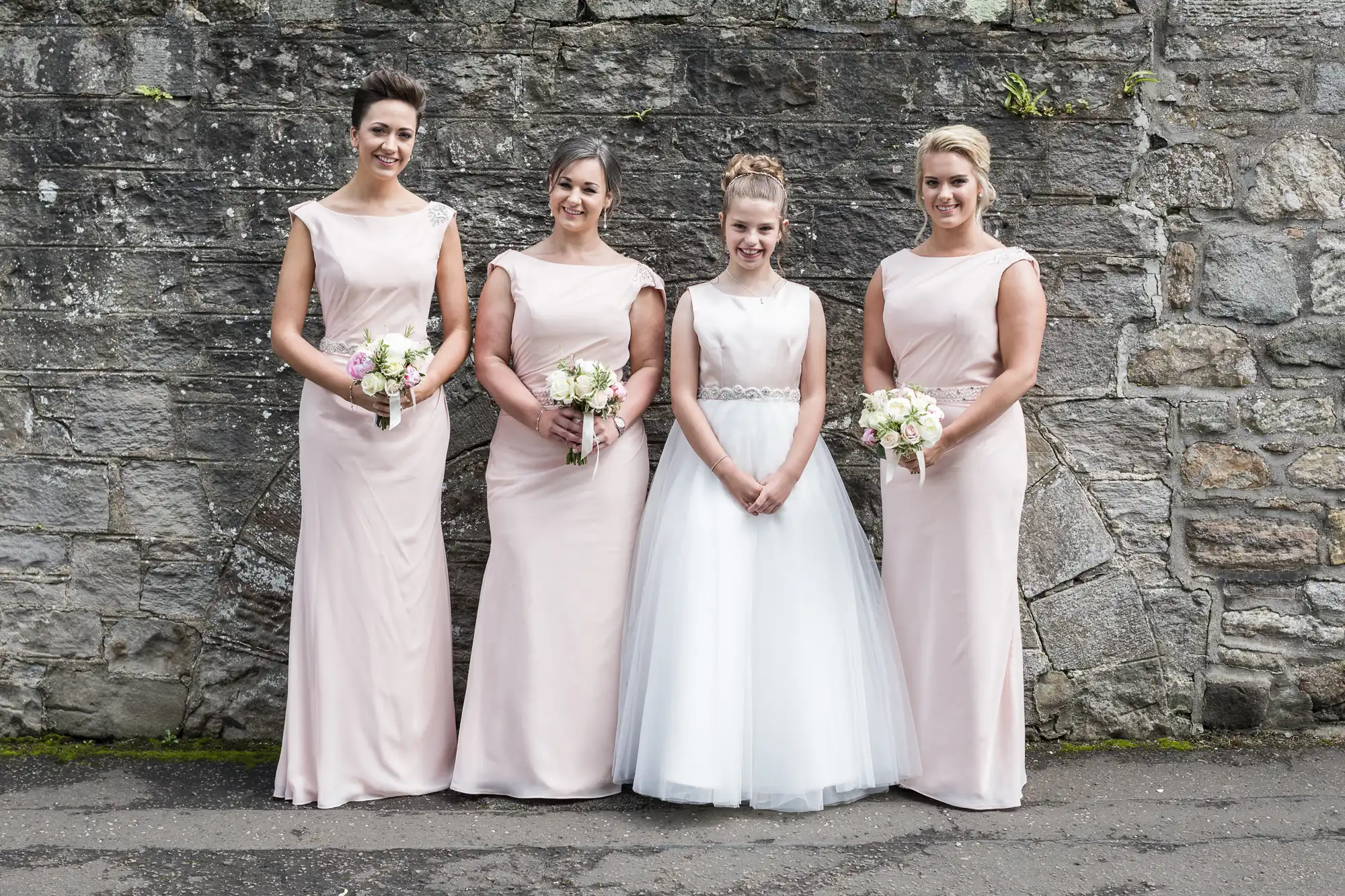 Four women are standing in front of a stone wall; three are wearing light pink dresses holding bouquets, and one, in the center, is wearing a white dress and holding her hands clasped.