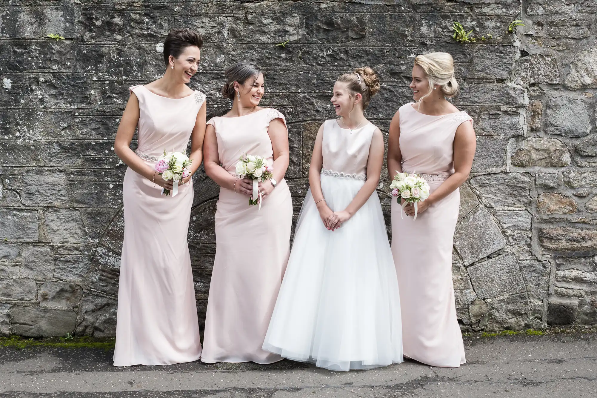 Four women in light pink dresses and a girl in a white dress stand in a row, holding bouquets and smiling, posing against a stone wall background.