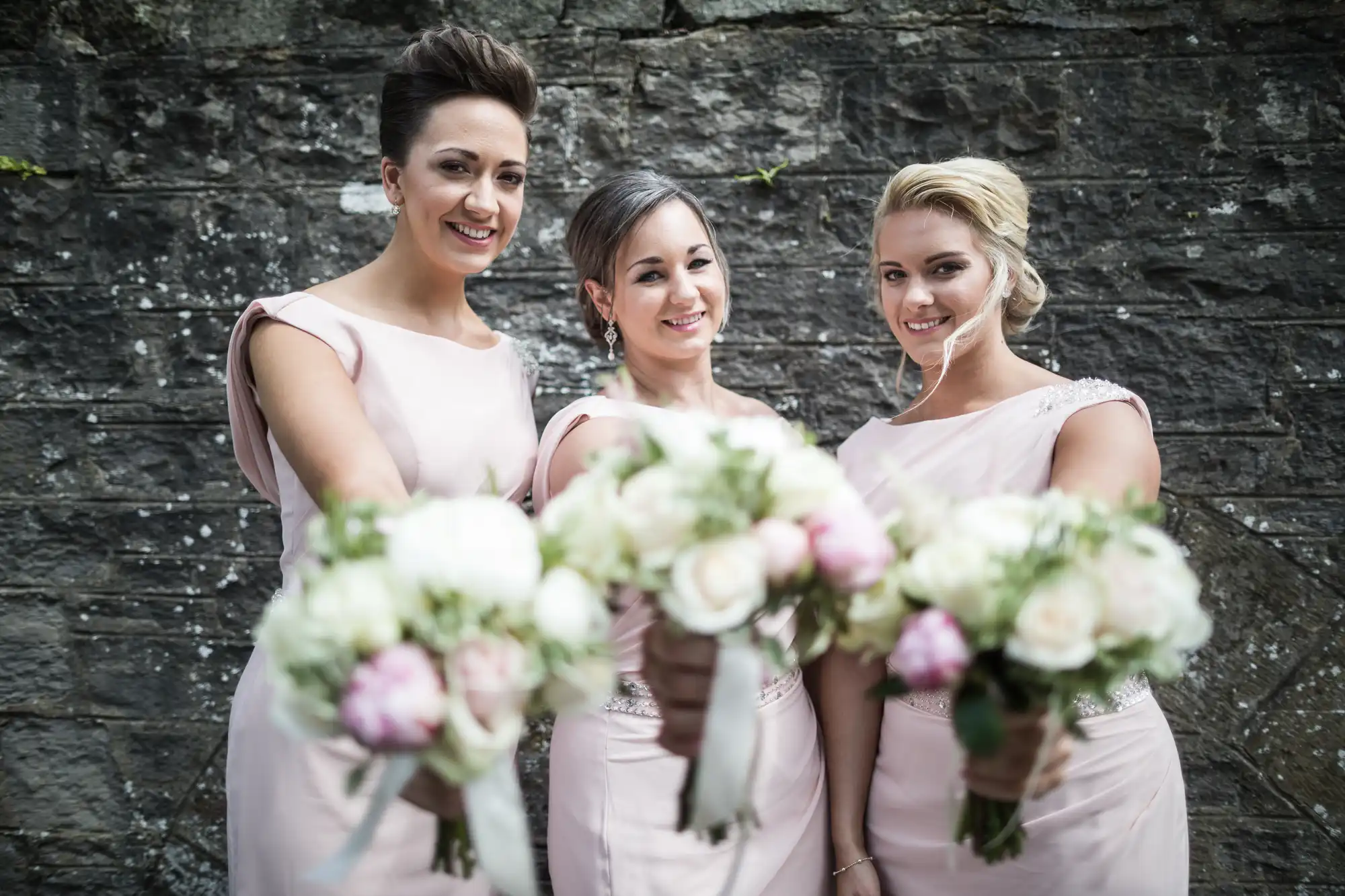 Three women in matching light pink dresses holding floral bouquets, standing in front of a stone wall.