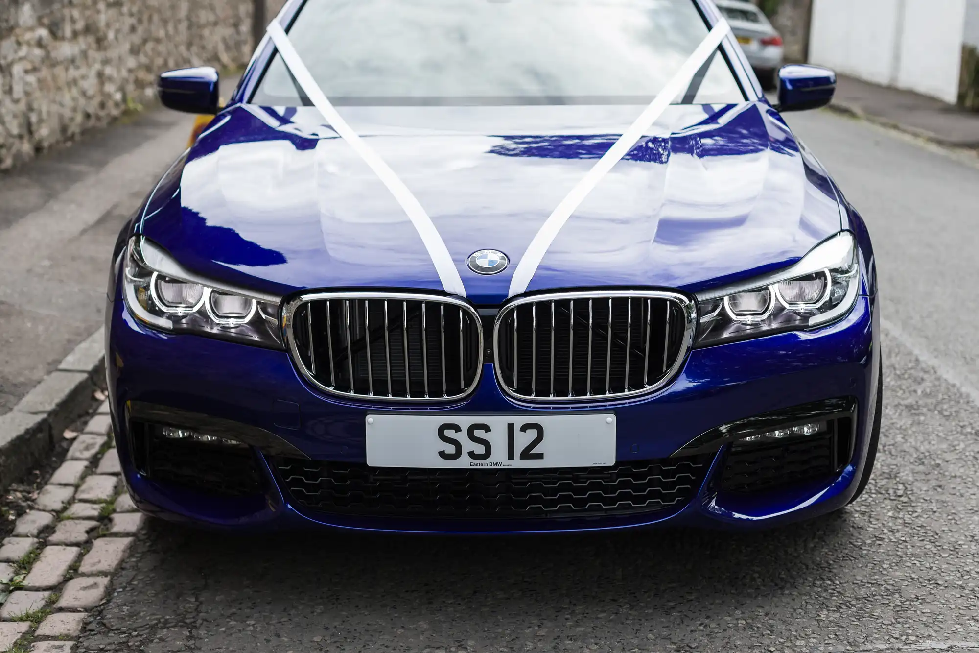 A blue car with a white ribbon on the hood is parked on the street. The license plate reads "SS 12.