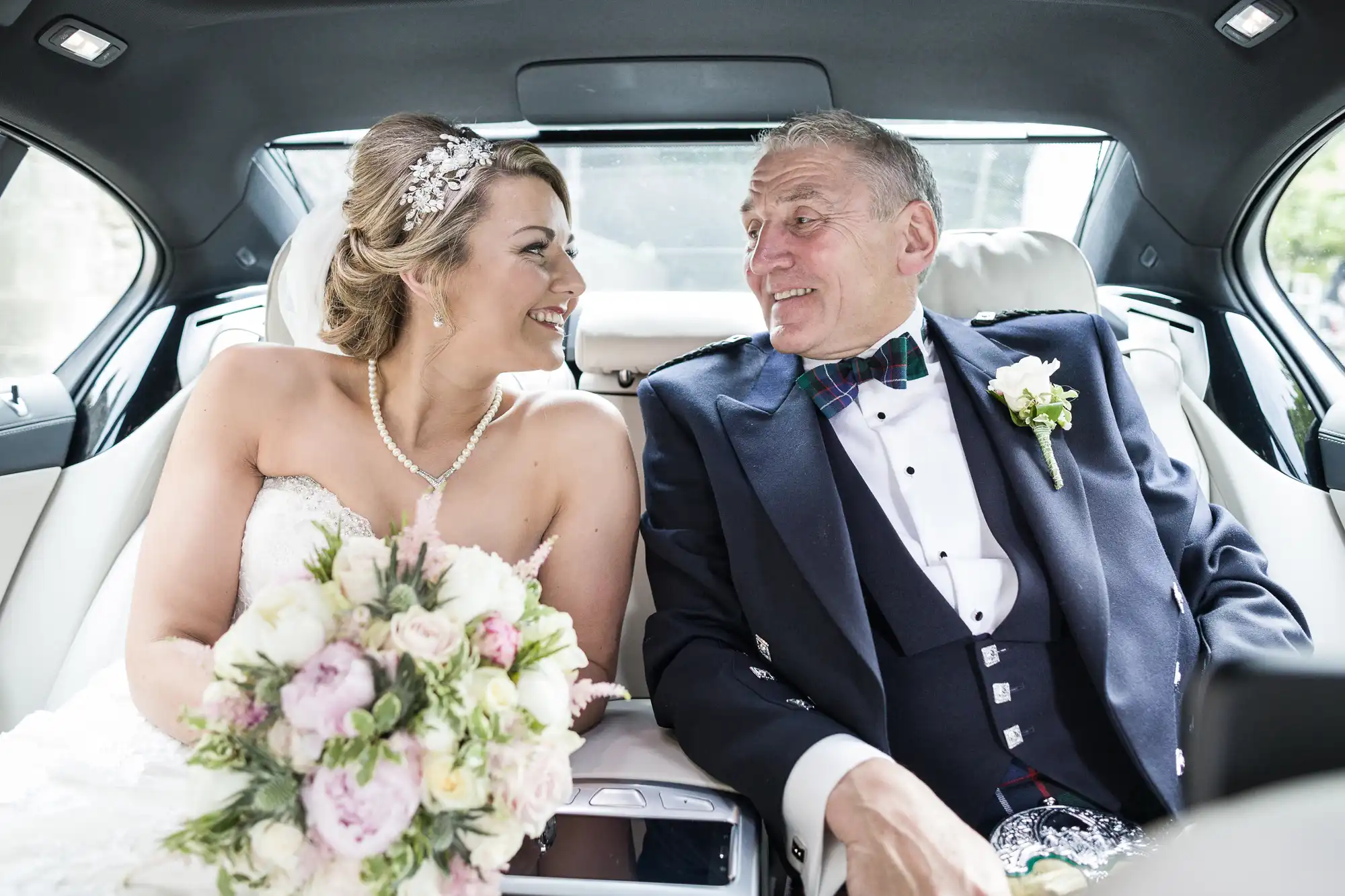 A smiling bride holding a bouquet and an older man in a suit, likely her father, share a moment in the backseat of a car.