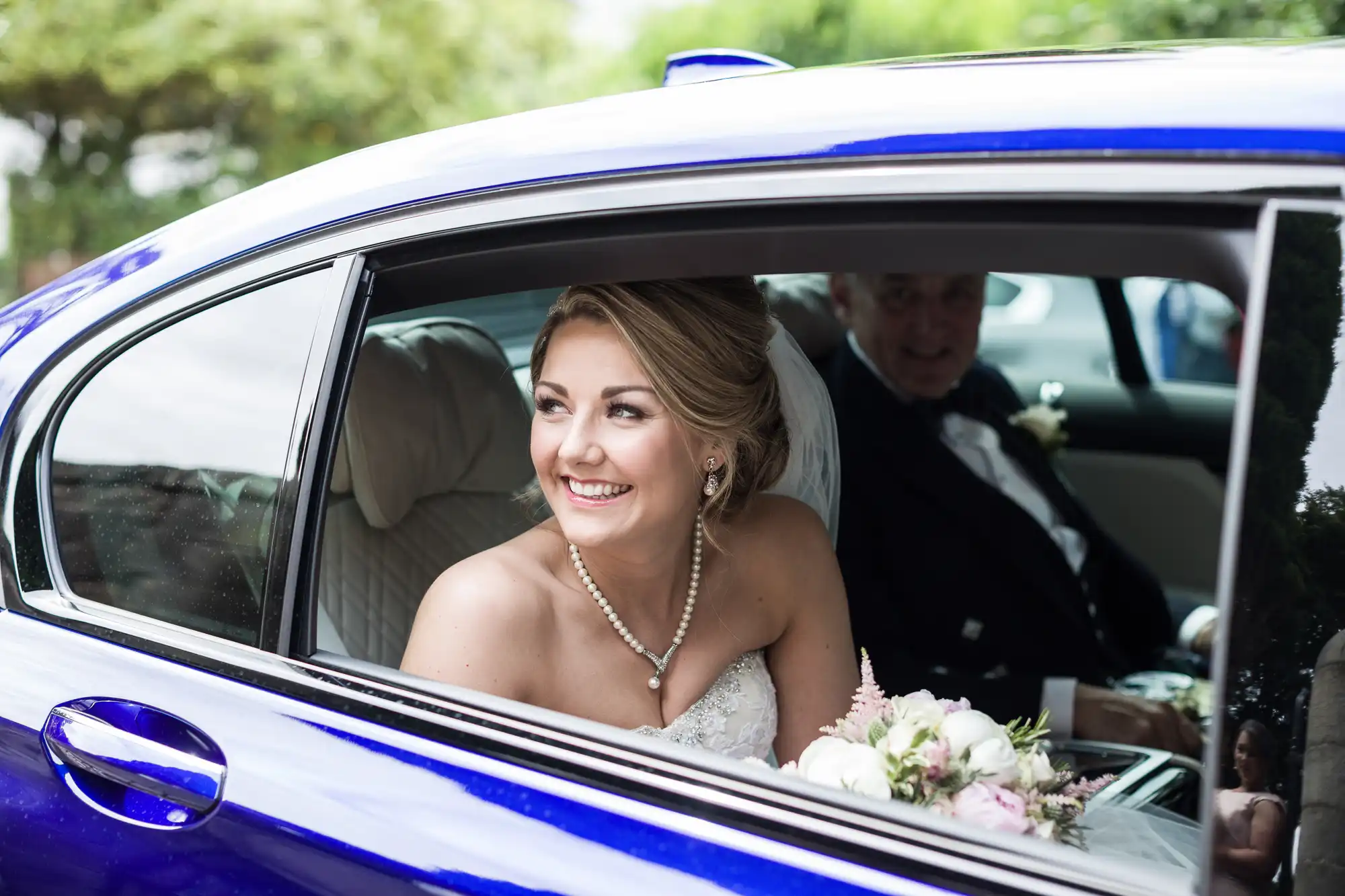 A smiling bride with a bouquet sits in the back seat of a blue car, looking out the open window. An older man in a tuxedo is seated next to her.