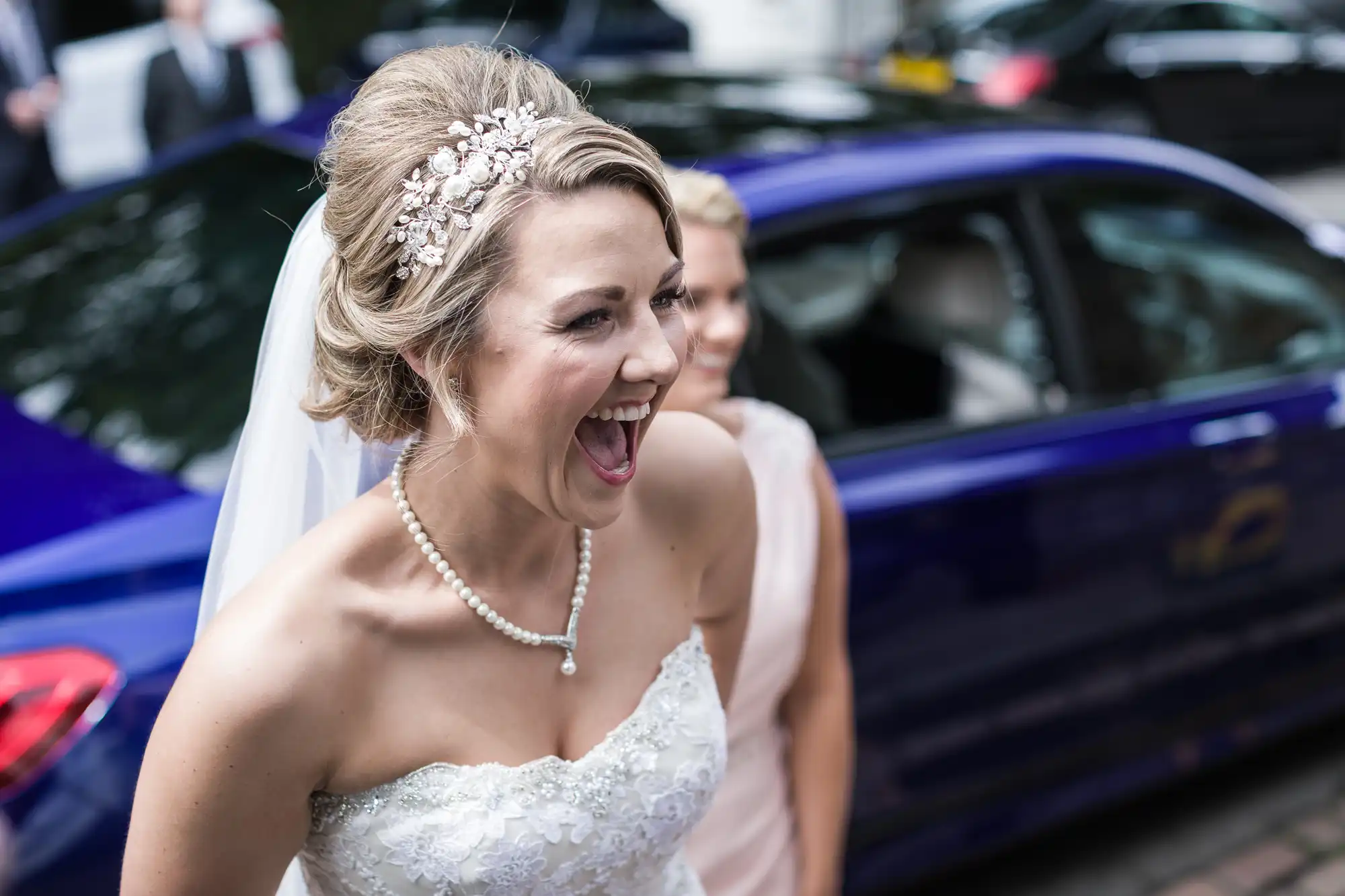 A smiling bride in a white strapless wedding dress and pearl necklace, adorned with a floral hairpiece, stands outdoors with a blue car in the background. Another woman in a pale dress is partially visible.