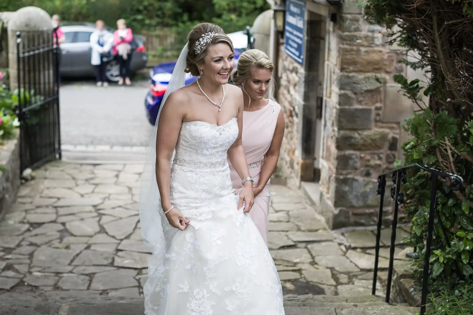 A bride in a white dress smiles as a bridesmaid in a pink dress helps her with the gown's train, walking on a stone path towards a building. Guests and parked vehicles are visible in the background.