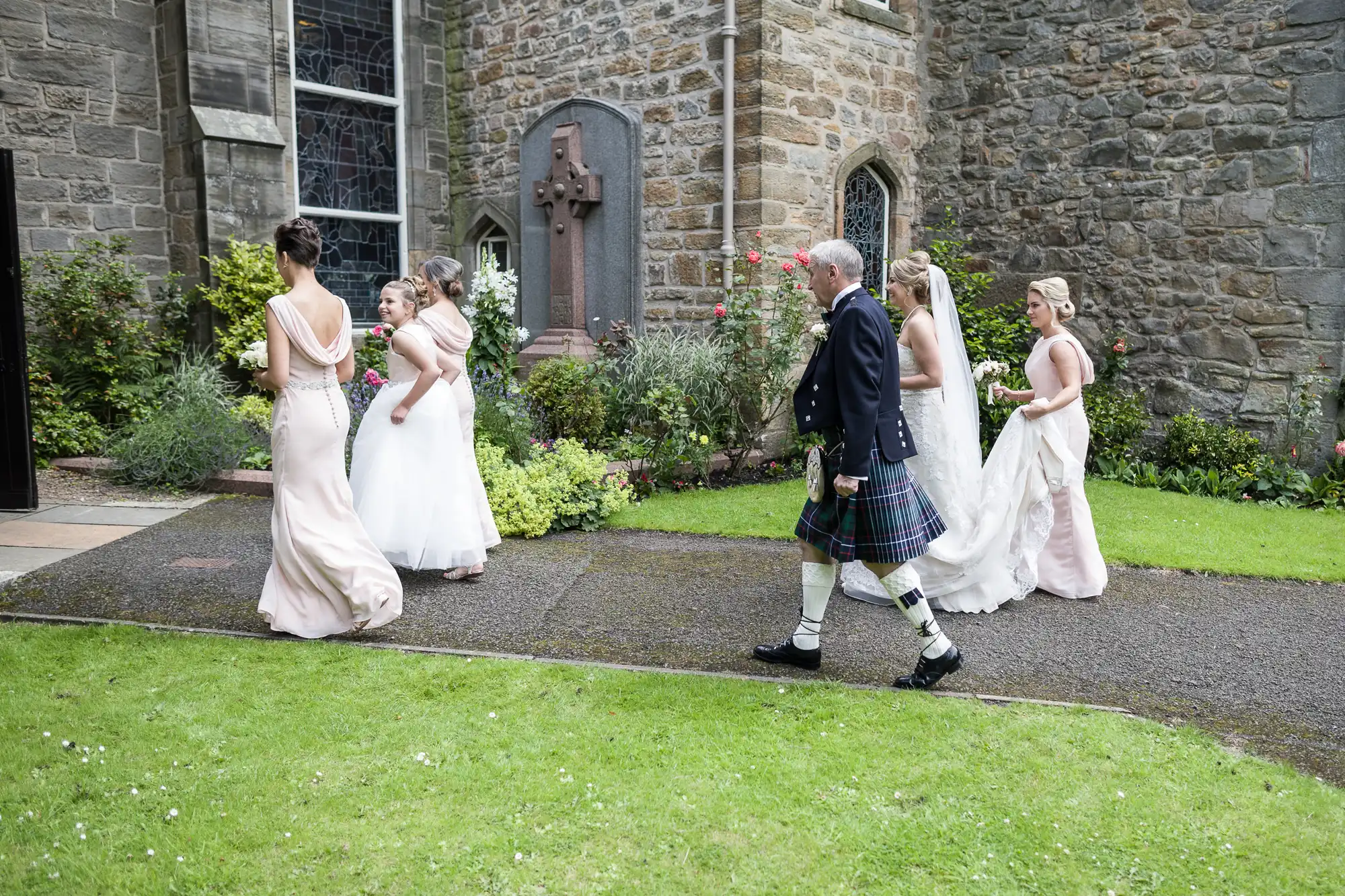 A group of women in formal dresses walk alongside a man in a kilt outside a stone building with stained glass windows and a cross monument, surrounded by greenery.