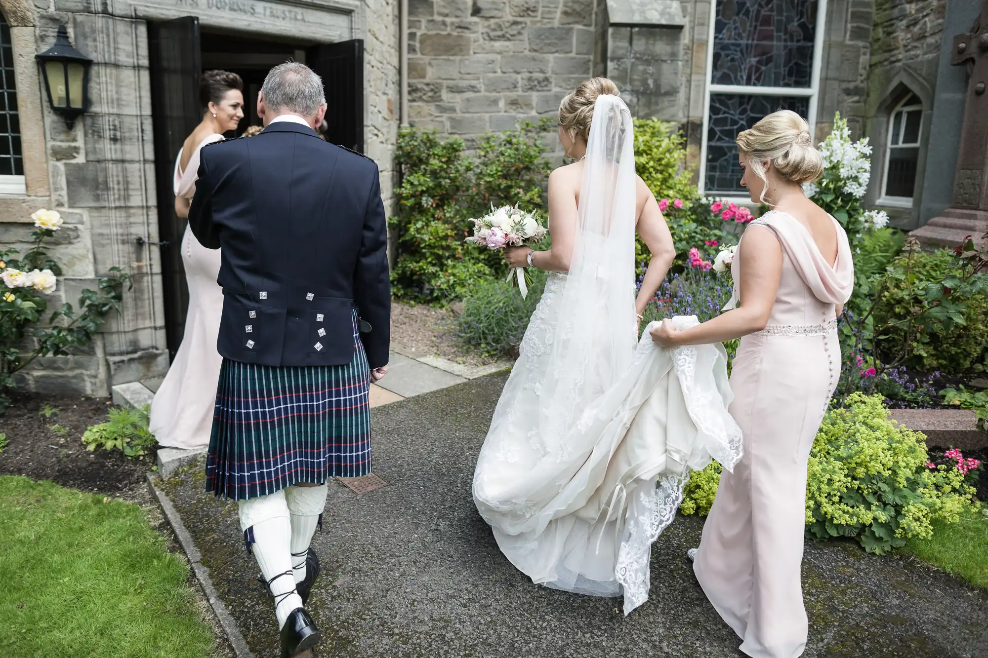 A bride in a white gown walks towards a stone church, accompanied by three others, one in a kilt and two in light dresses, one of whom holds the bride's dress.