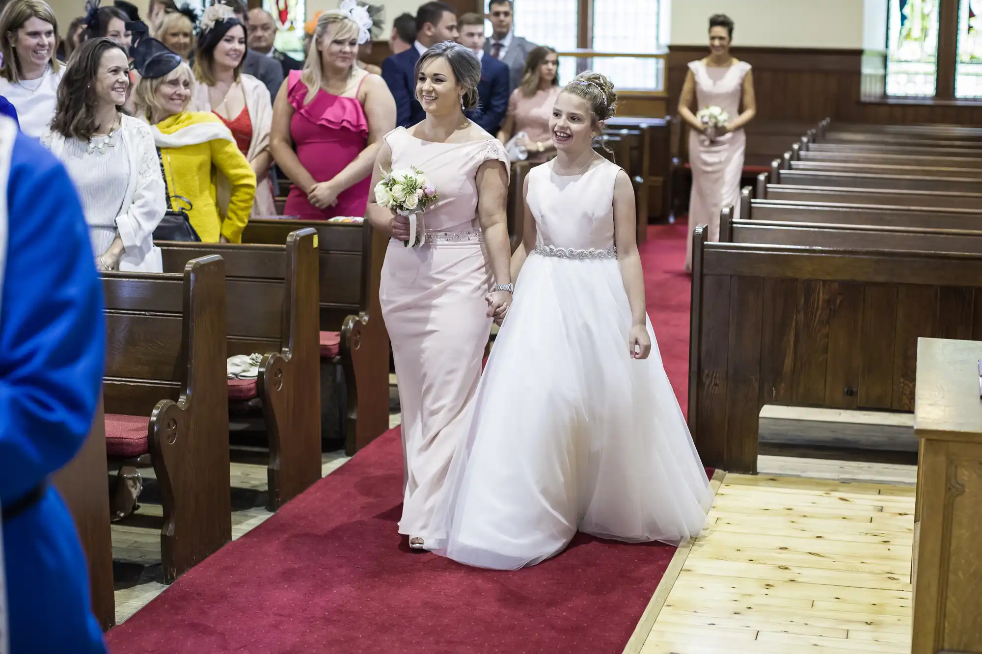 Two women, dressed in formal gowns, walk down the aisle in a church, smiling. One holds a bouquet. Guests, seated in pews, watch the procession.