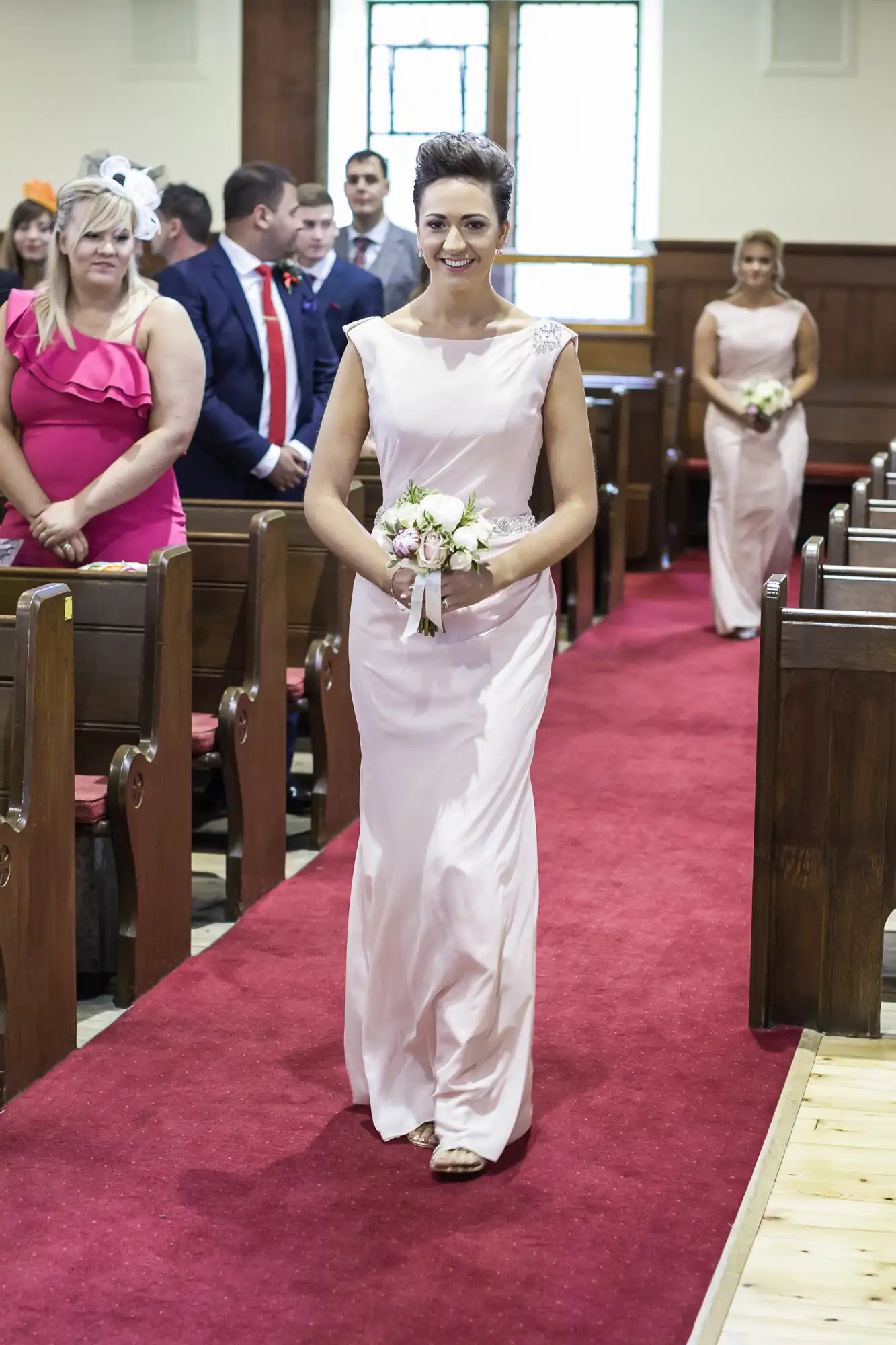 A woman in a light pink dress walks down a red carpet aisle holding a bouquet, with people seated on wooden benches on either side, in a church setting.