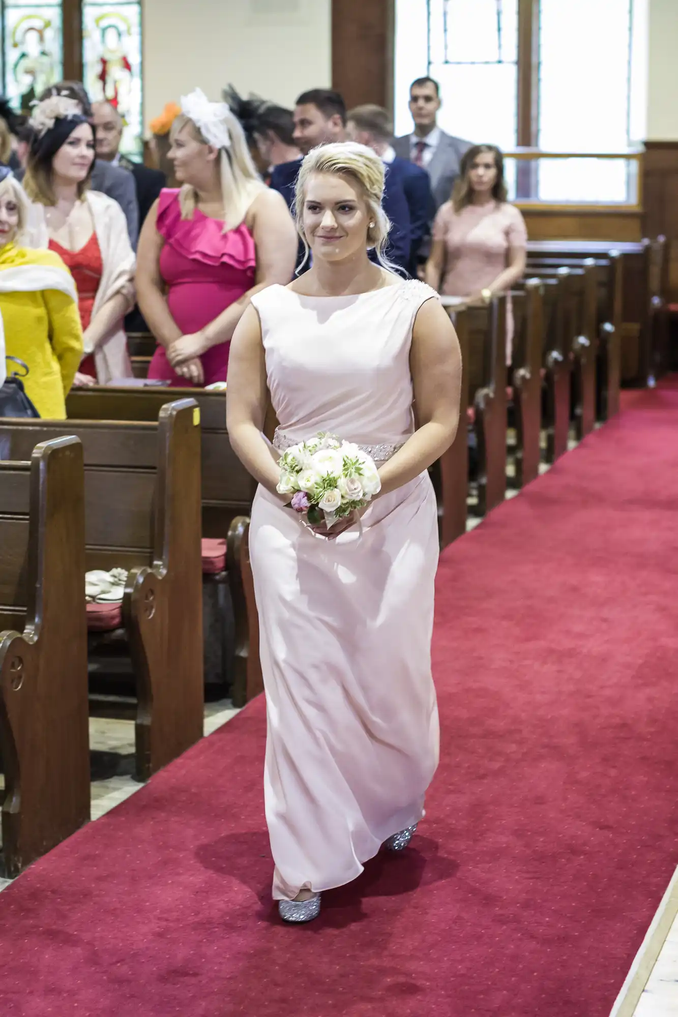 A woman in a light pink dress walks down a red carpeted aisle in a church, holding a small bouquet of flowers, with seated guests watching.