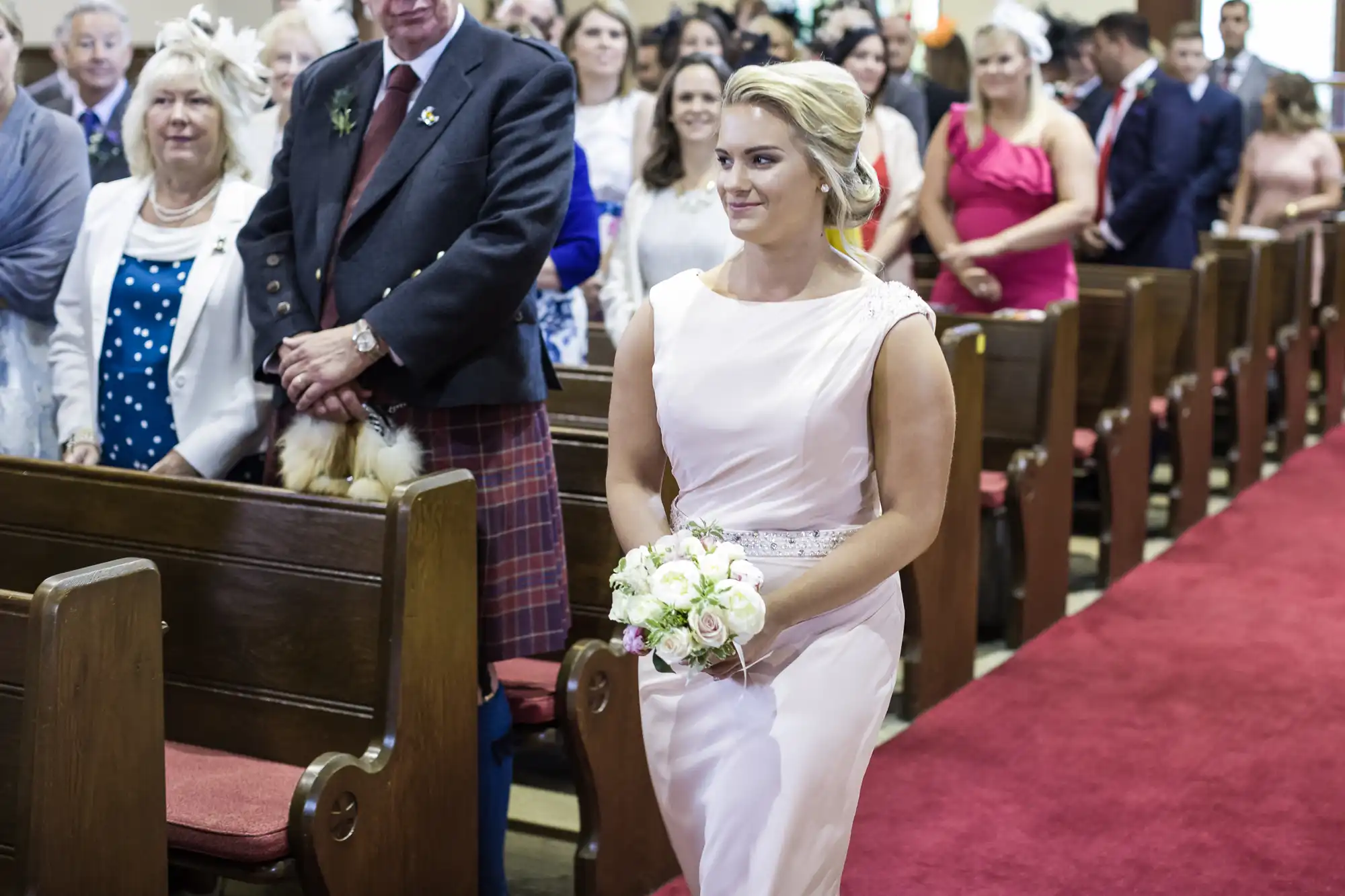 A woman in a light pink dress holds a bouquet of flowers and walks down the aisle in a church, with seated guests looking on.