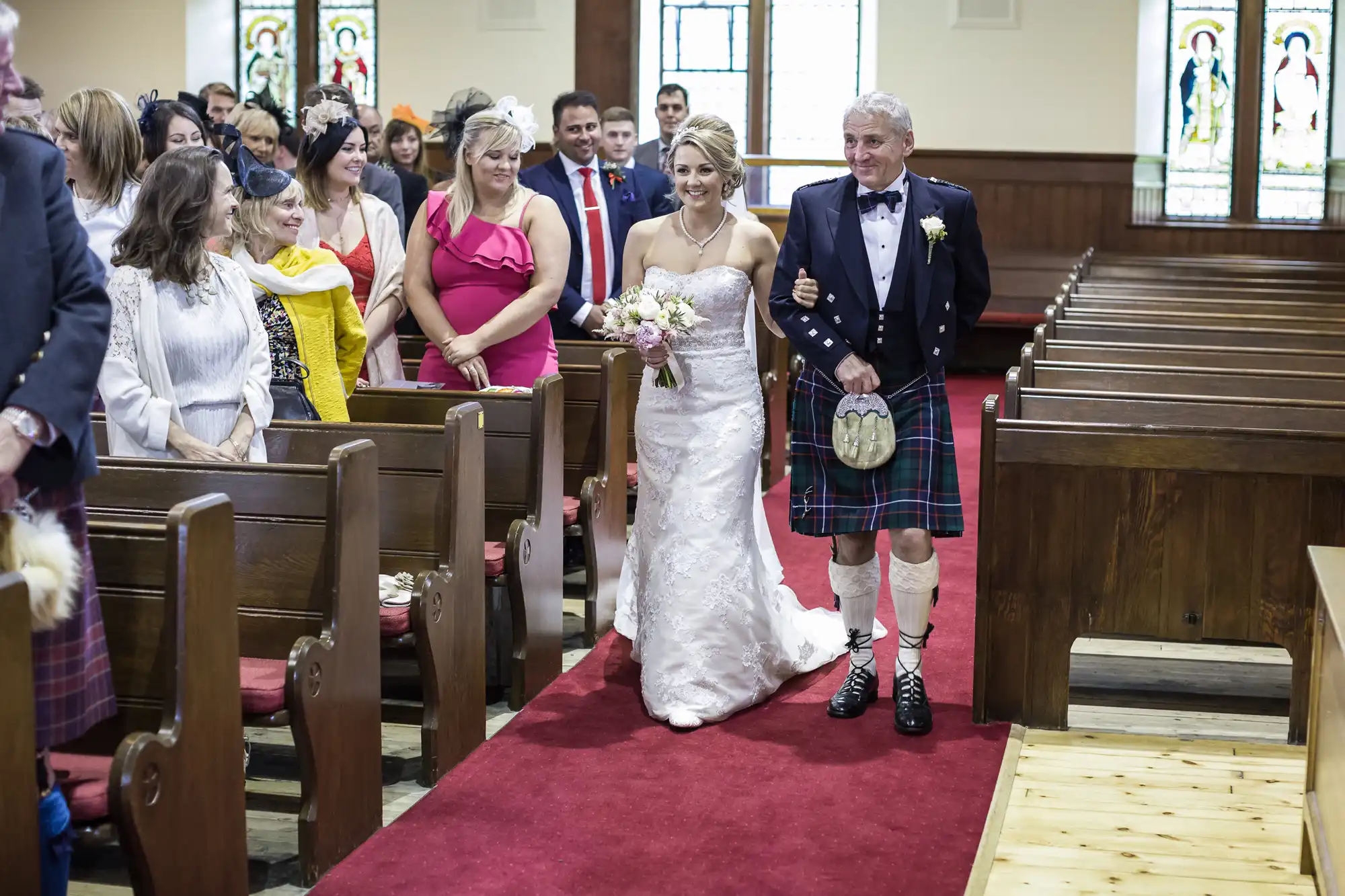 A bride in a white gown walks down the aisle with an older man in a kilt in a church, followed by guests seated in pews.