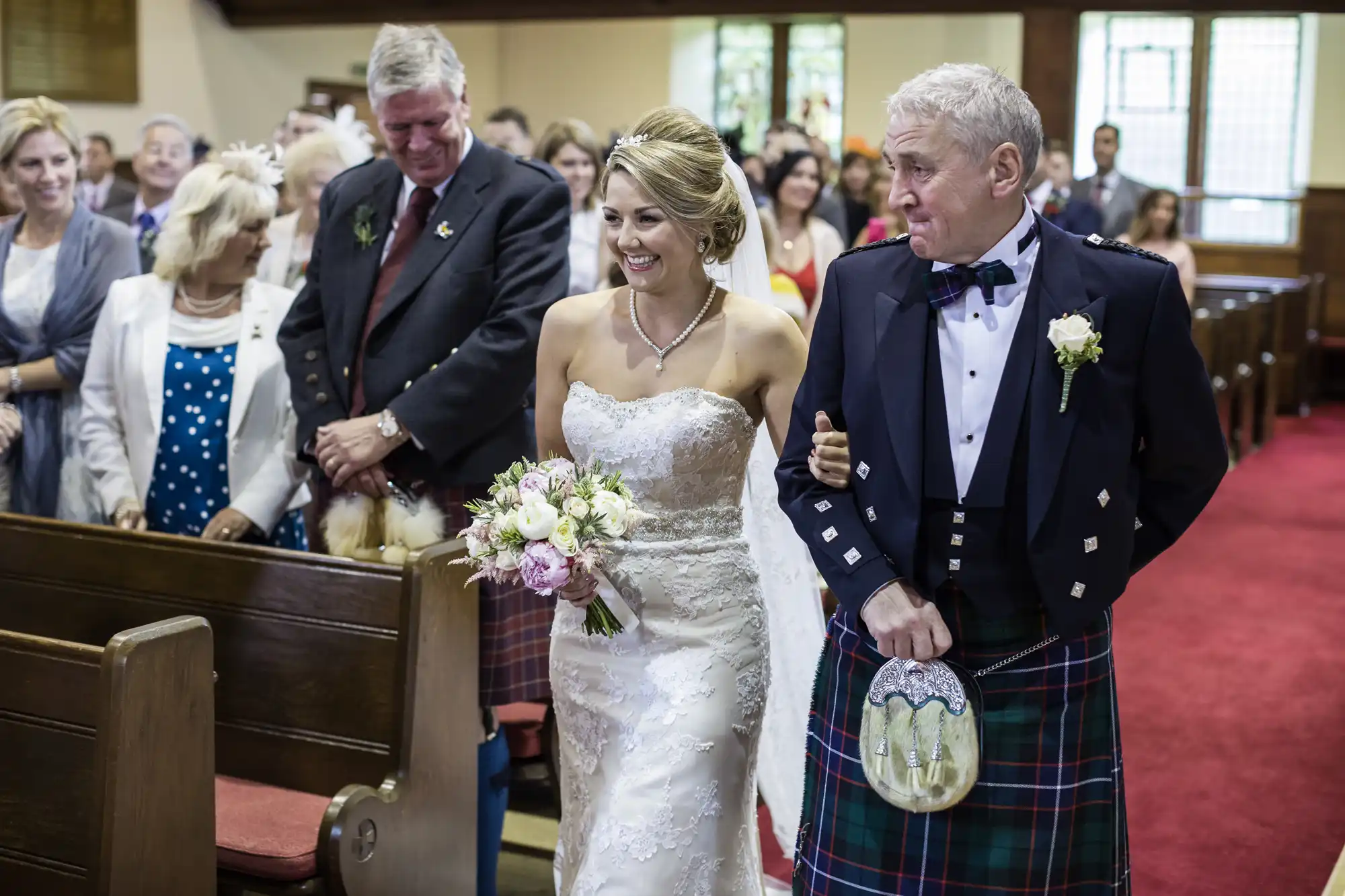 A bride in a white dress, holding flowers, walks down the aisle with an older man in a kilt and formal attire, while guests watch in a church setting.