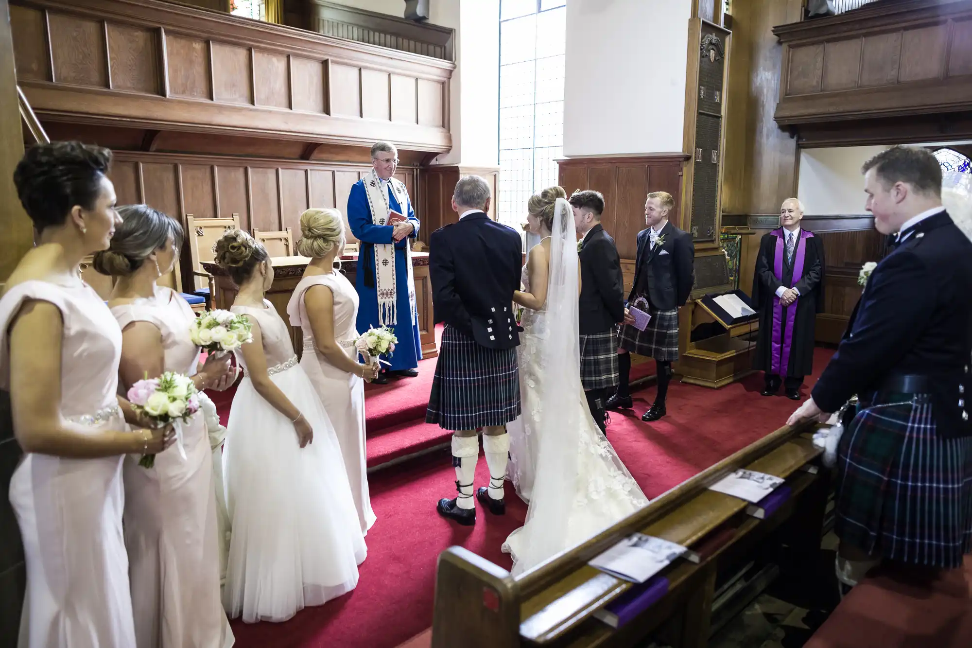 A wedding ceremony in a church with a bride and groom standing before the officiant. Bridesmaids in pale dresses and groomsmen in kilts are positioned nearby.