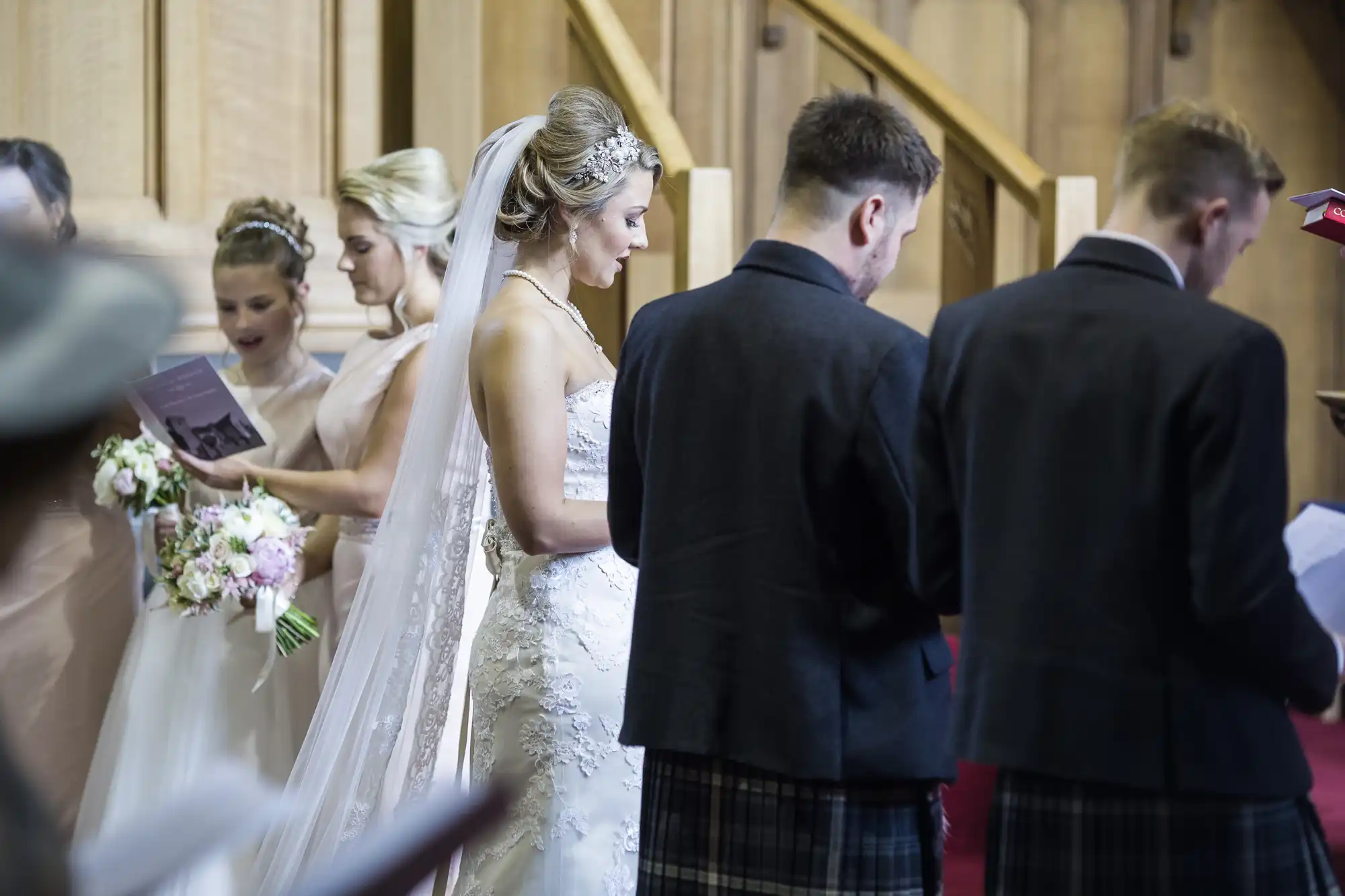 A bride and bridesmaids stand holding bouquets and papers, while two men in kilts stand nearby with their backs to the camera inside a wooden-paneled venue.