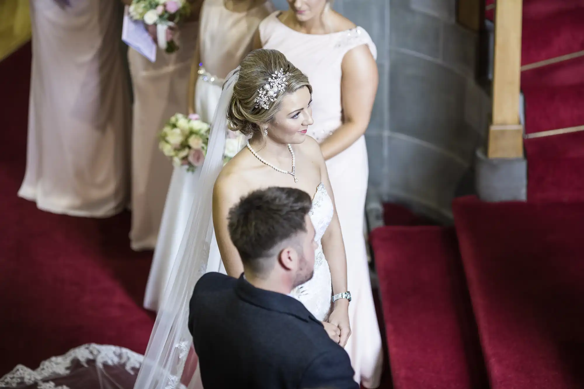 A bride in a white gown with a long veil stands beside a groom in a dark suit on red-carpeted steps, accompanied by bridesmaids holding floral bouquets.