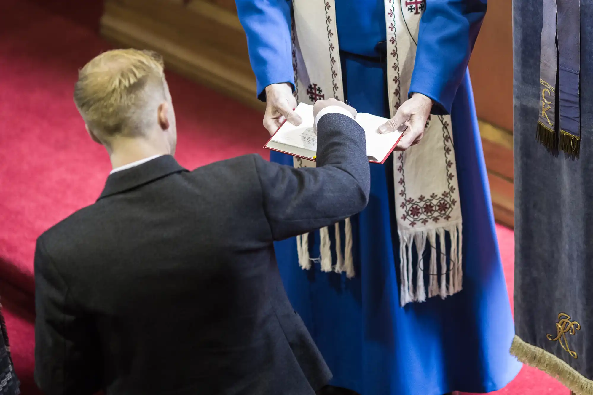 A kneeling person in a suit extends a hand to touch a book held by another person in a blue robe, who is standing.