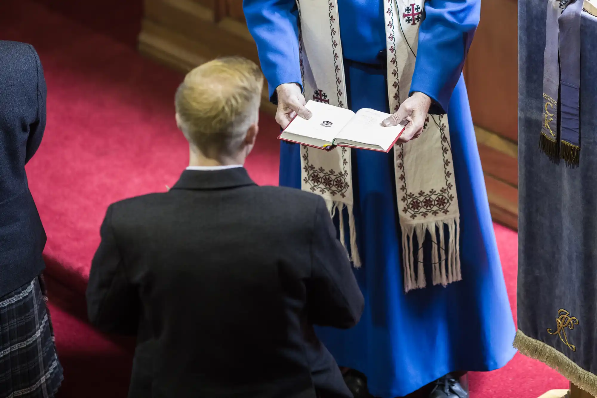 A person kneels in front of a religious figure holding an open book, possibly a Bible, during a ceremony in a church setting.