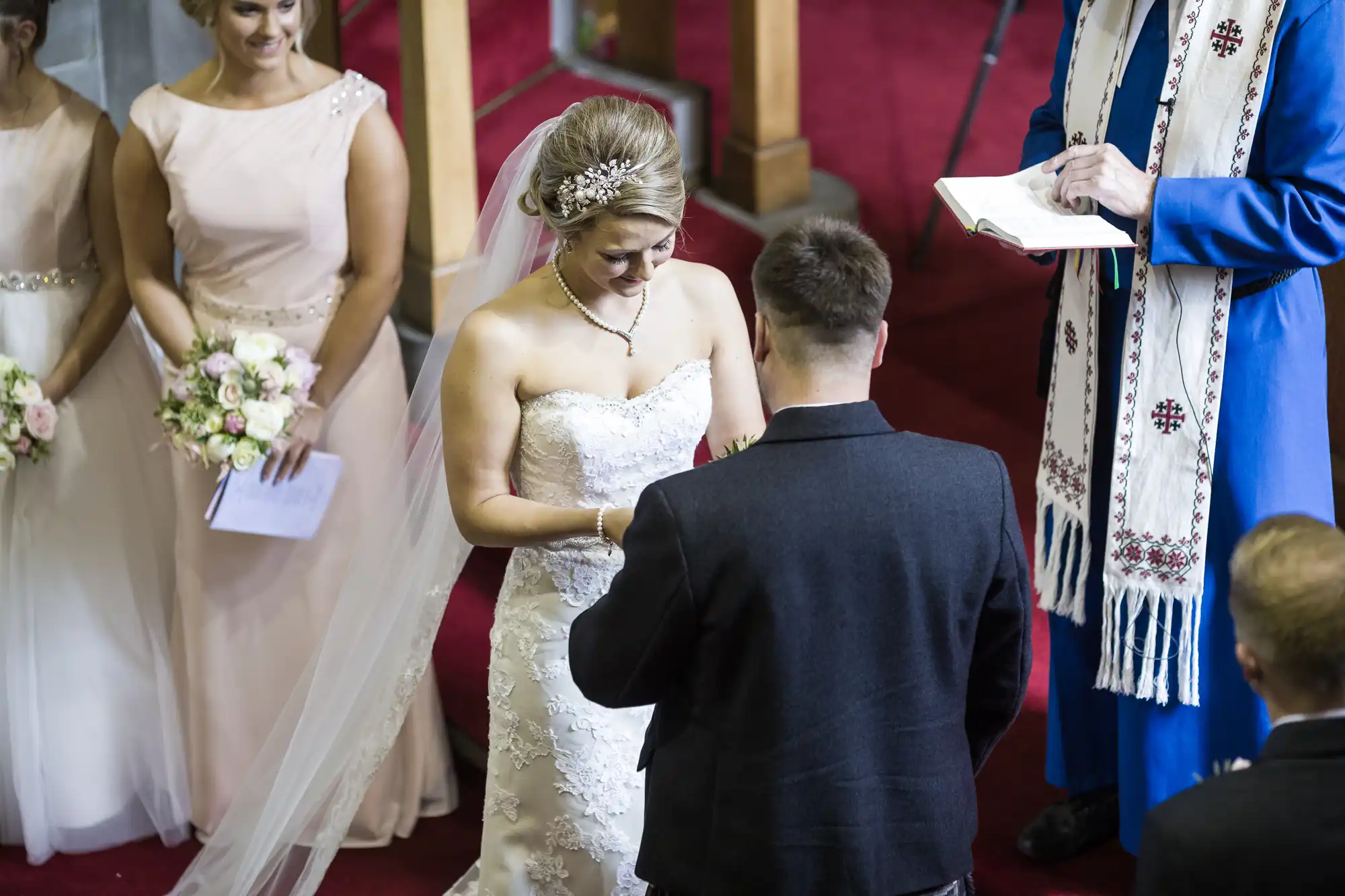 A bride and groom exchange rings during their wedding ceremony in a church. The bride's attendants stand nearby, holding bouquets.