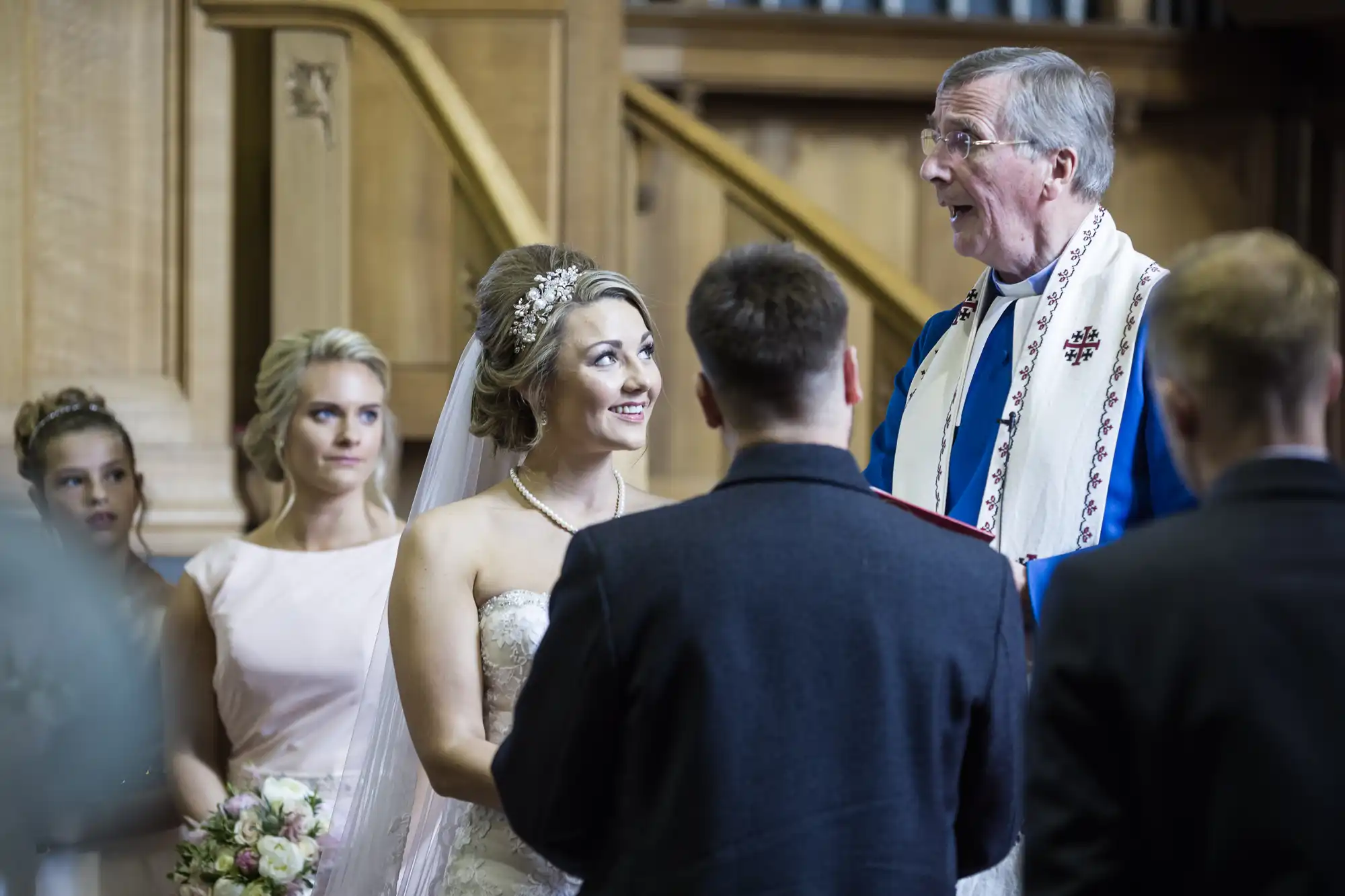 A couple stands before a clergyman at a wedding ceremony. The bride, holding a bouquet, is smiling while the groom stands with his back to the camera. Two bridesmaids are visible in the background.