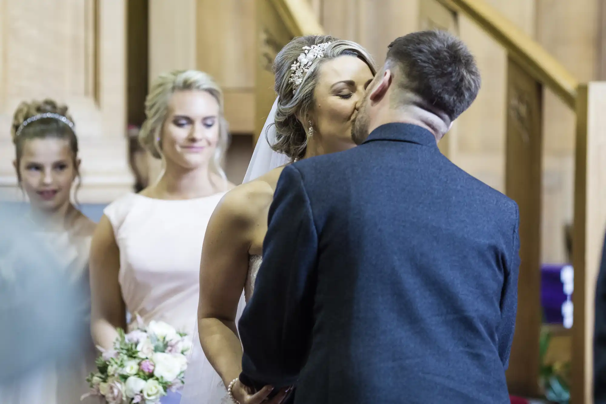 A bride and groom share a kiss at their wedding ceremony. Bridesmaids stand in the background, one holding a bouquet of flowers.