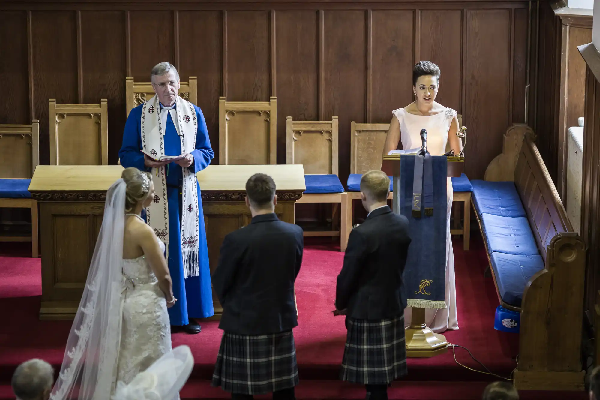 A couple stands before a clergyman and a speaker during their wedding ceremony in a church. The groom and groomsman wear kilts, while the bride wears a white gown with a long veil.