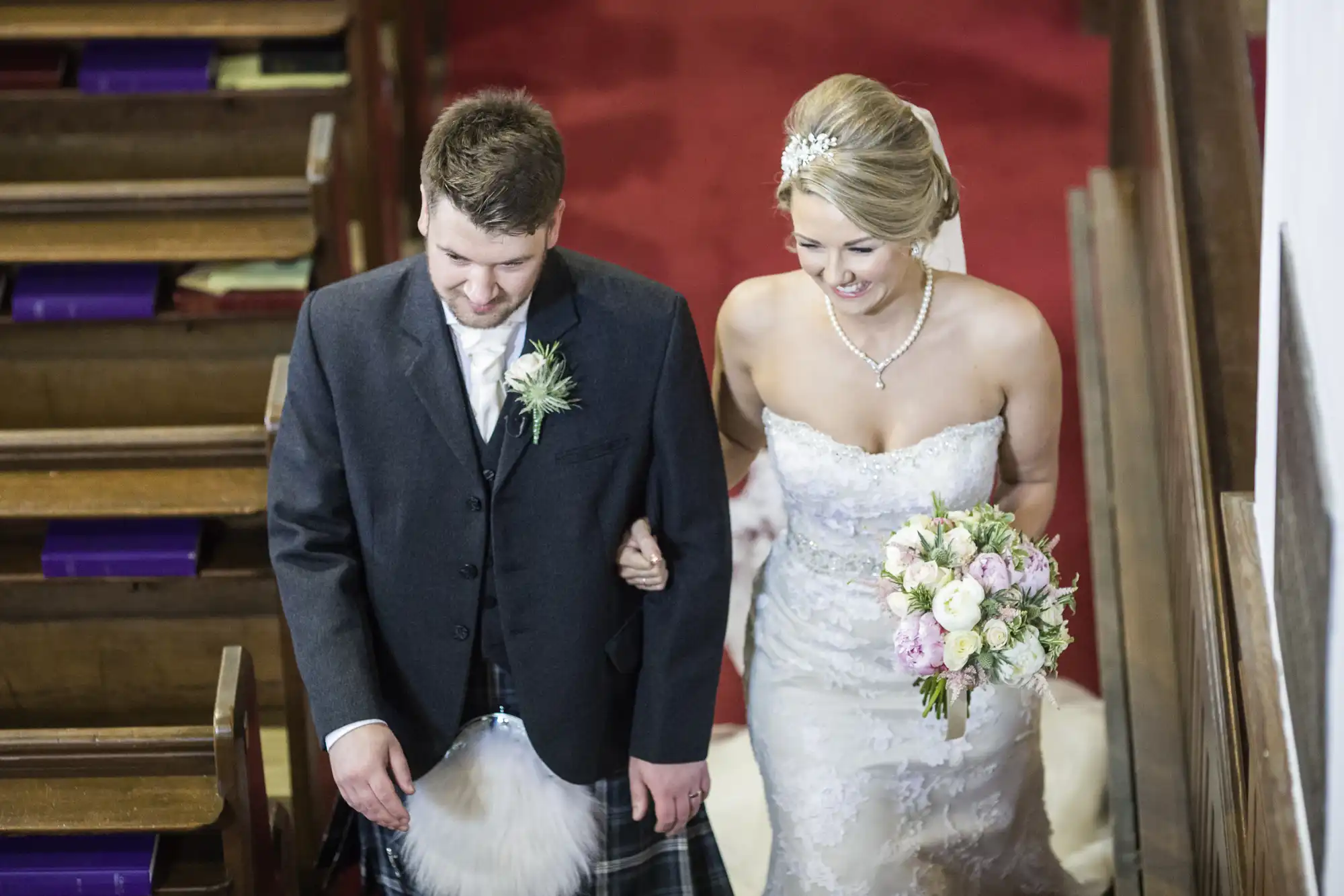 Bride and groom walk down the aisle. The groom wears traditional Scottish attire, and the bride is in a white strapless wedding dress holding a bouquet of flowers.