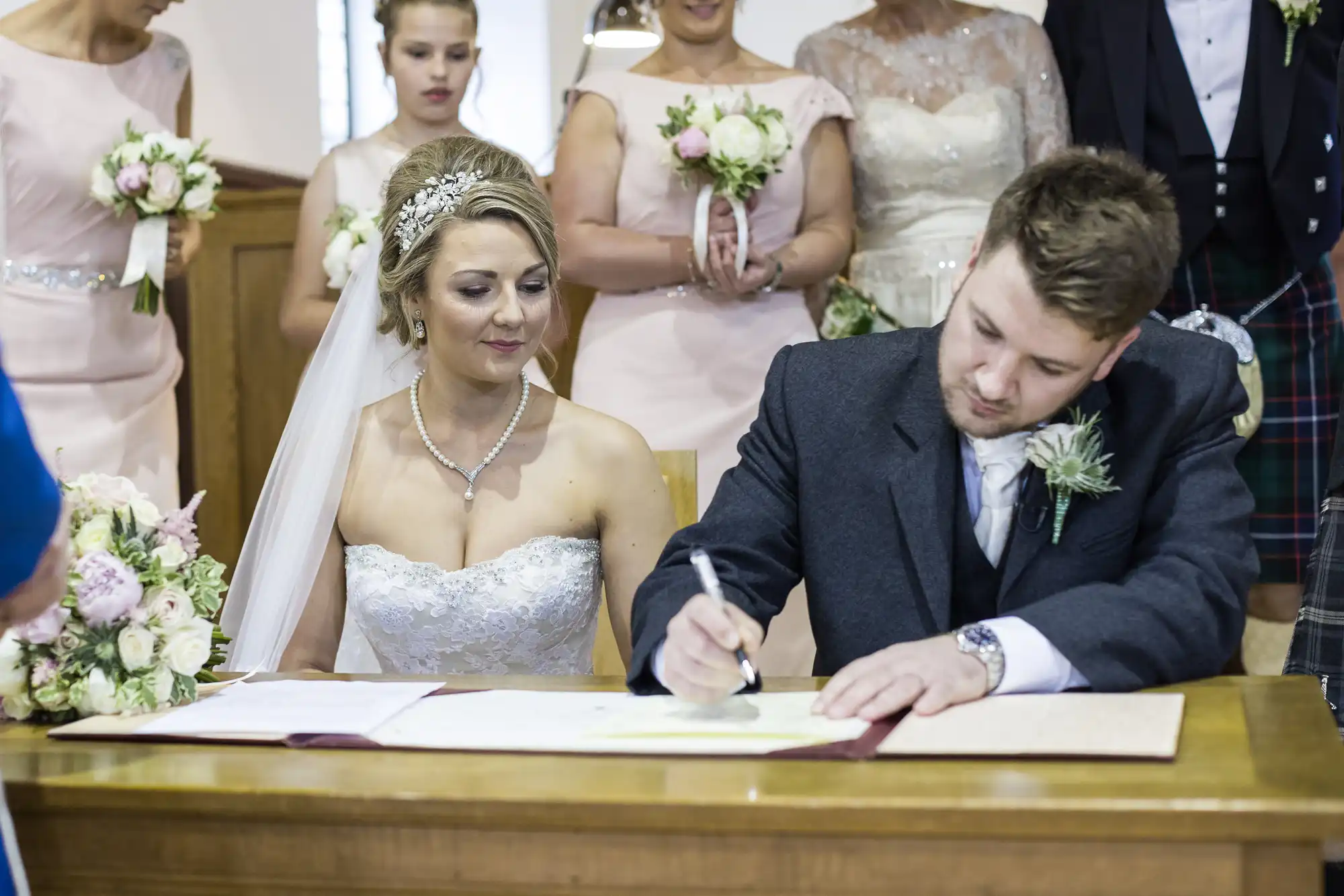 A bride in a white wedding dress and a groom in a suit sign a document at a table, surrounded by bridesmaids holding bouquets in a wedding ceremony.