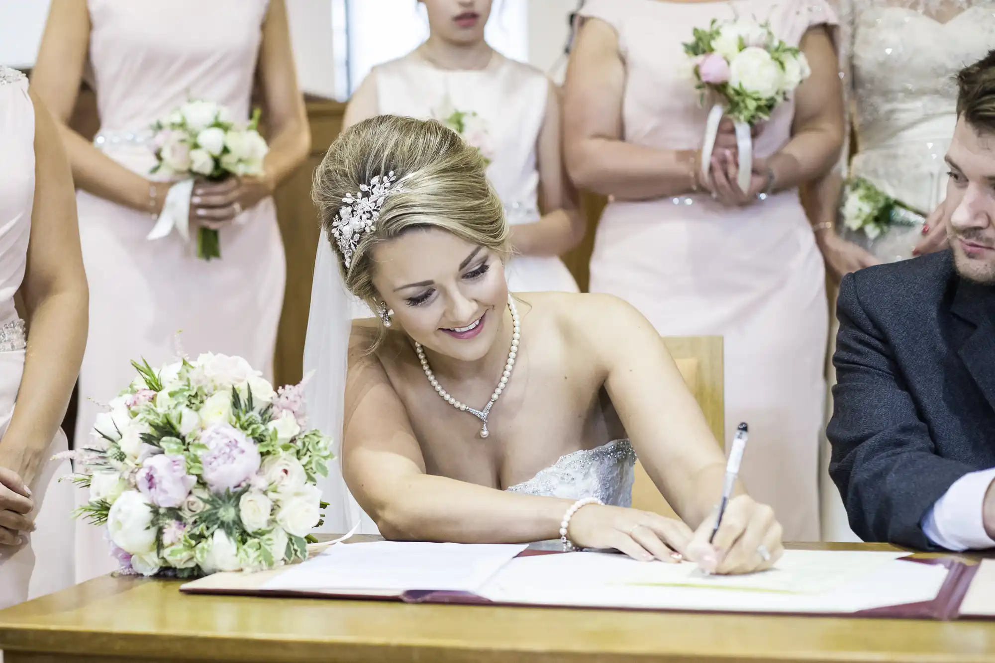 A bride in a white dress and tiara signs a document at a table, surrounded by bridesmaids in light pink dresses holding bouquets. A man in a suit sits beside her.