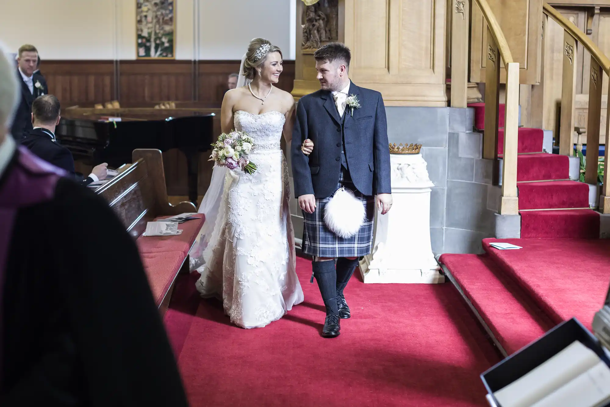 A bride in a white gown and a groom in a kilt walk arm in arm down a church aisle, smiling at each other.