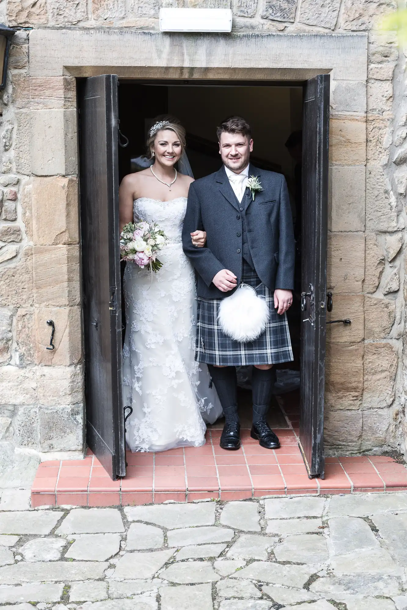 A bride in a white gown and a groom in a kilt and jacket stand arm in arm in a doorway, smiling. The bride holds a bouquet of flowers.