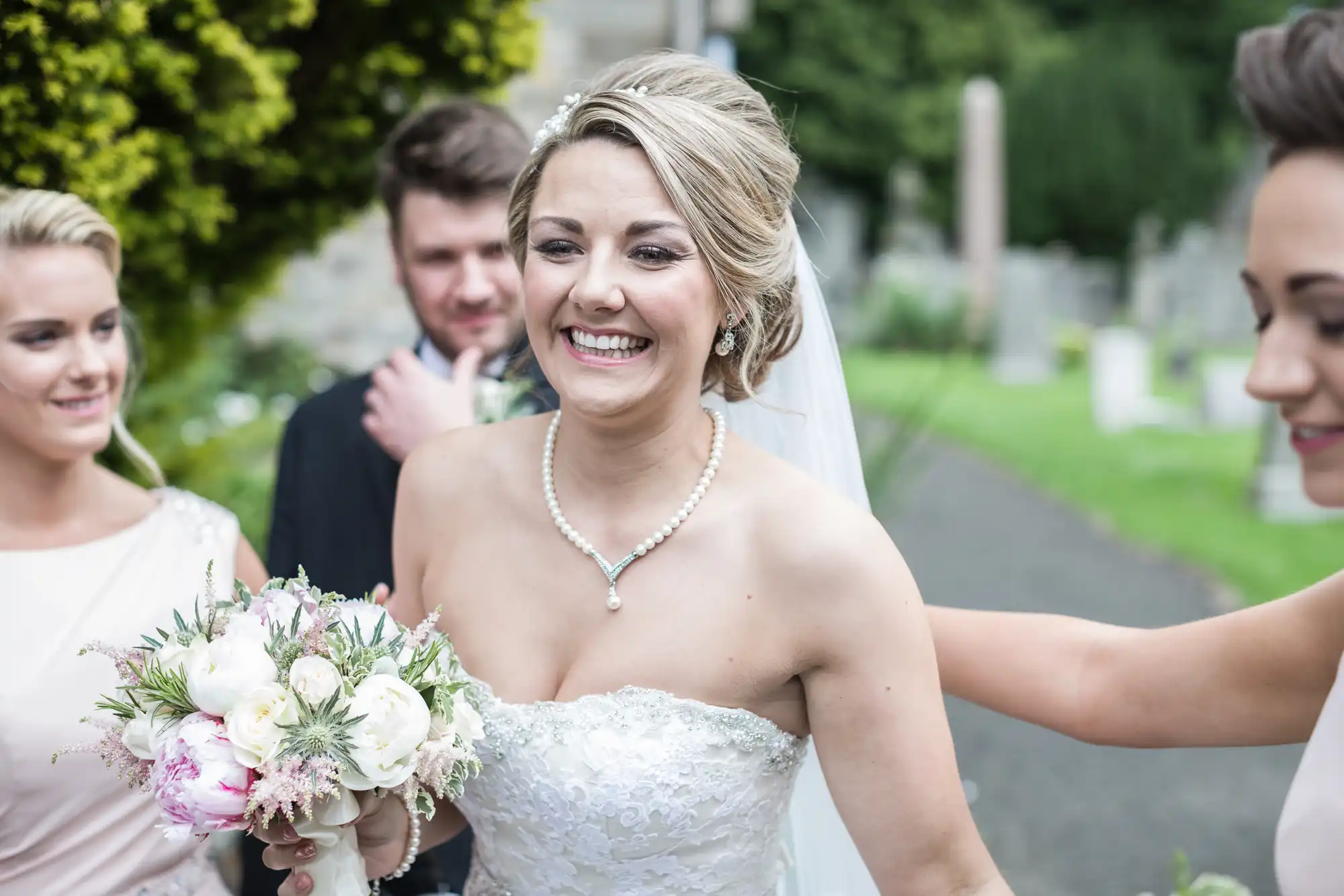 A bride in a white dress smiles while holding a bouquet of flowers, standing with three other people outdoors near greenery.