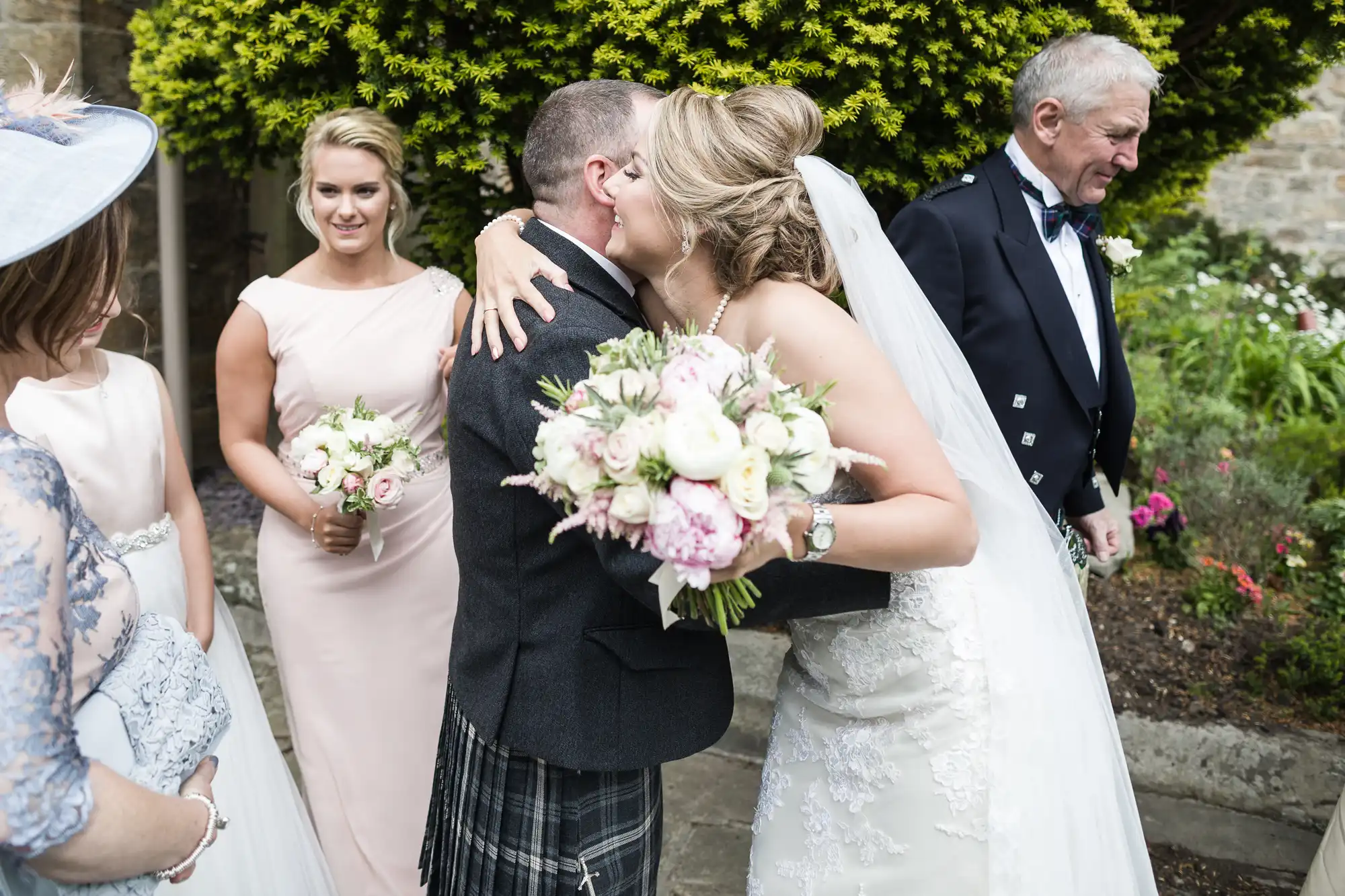 A bride, holding a bouquet and wearing a white dress, hugs a groom in a kilt. A bridesmaid and two other guests stand nearby. They are outdoors by a greenery backdrop.