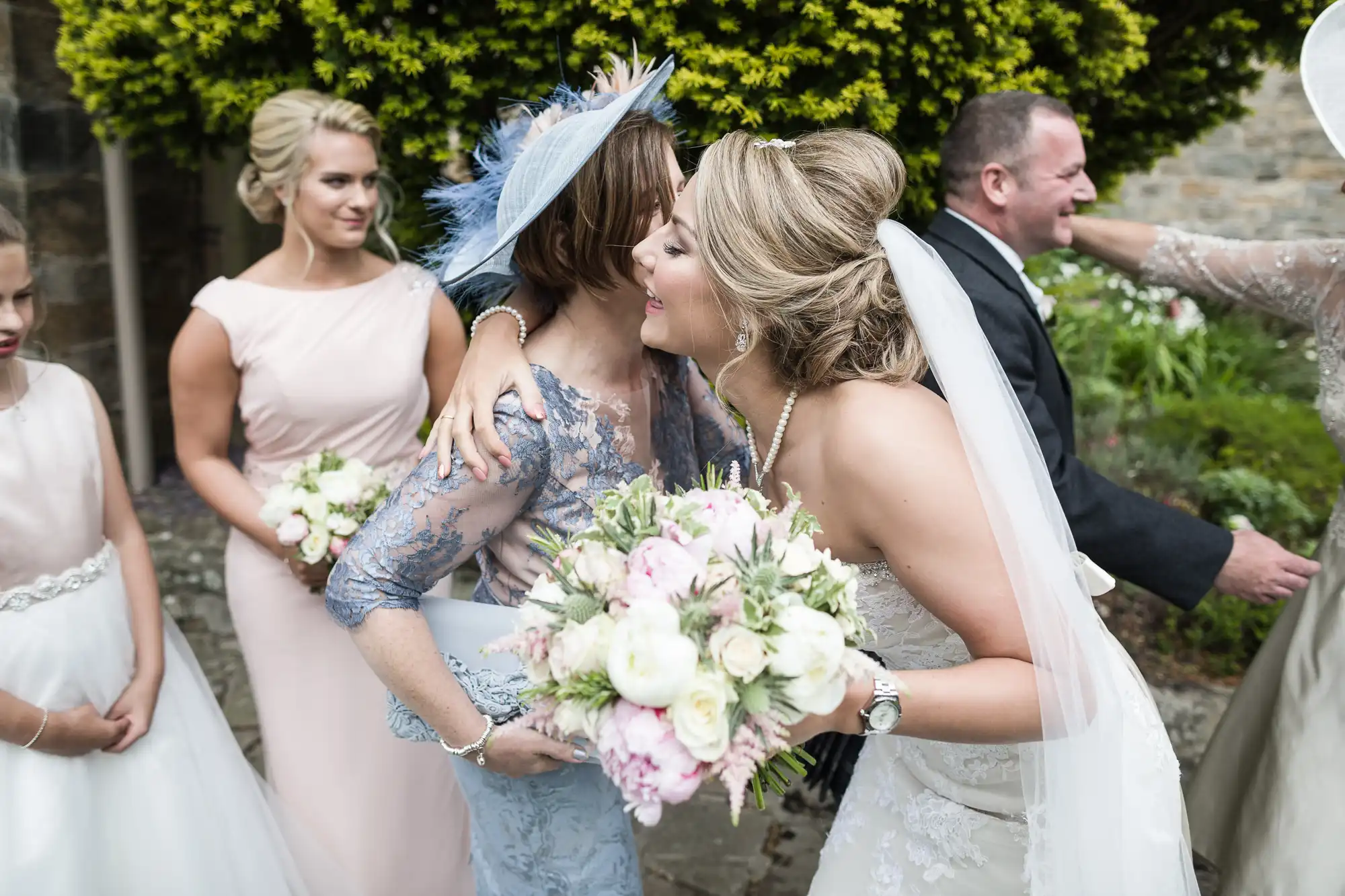 Bride in a white dress embraces a woman in a blue dress while holding a flower bouquet. A groom and bridesmaids are in the background, accompanied by flower girls in light pink dresses.