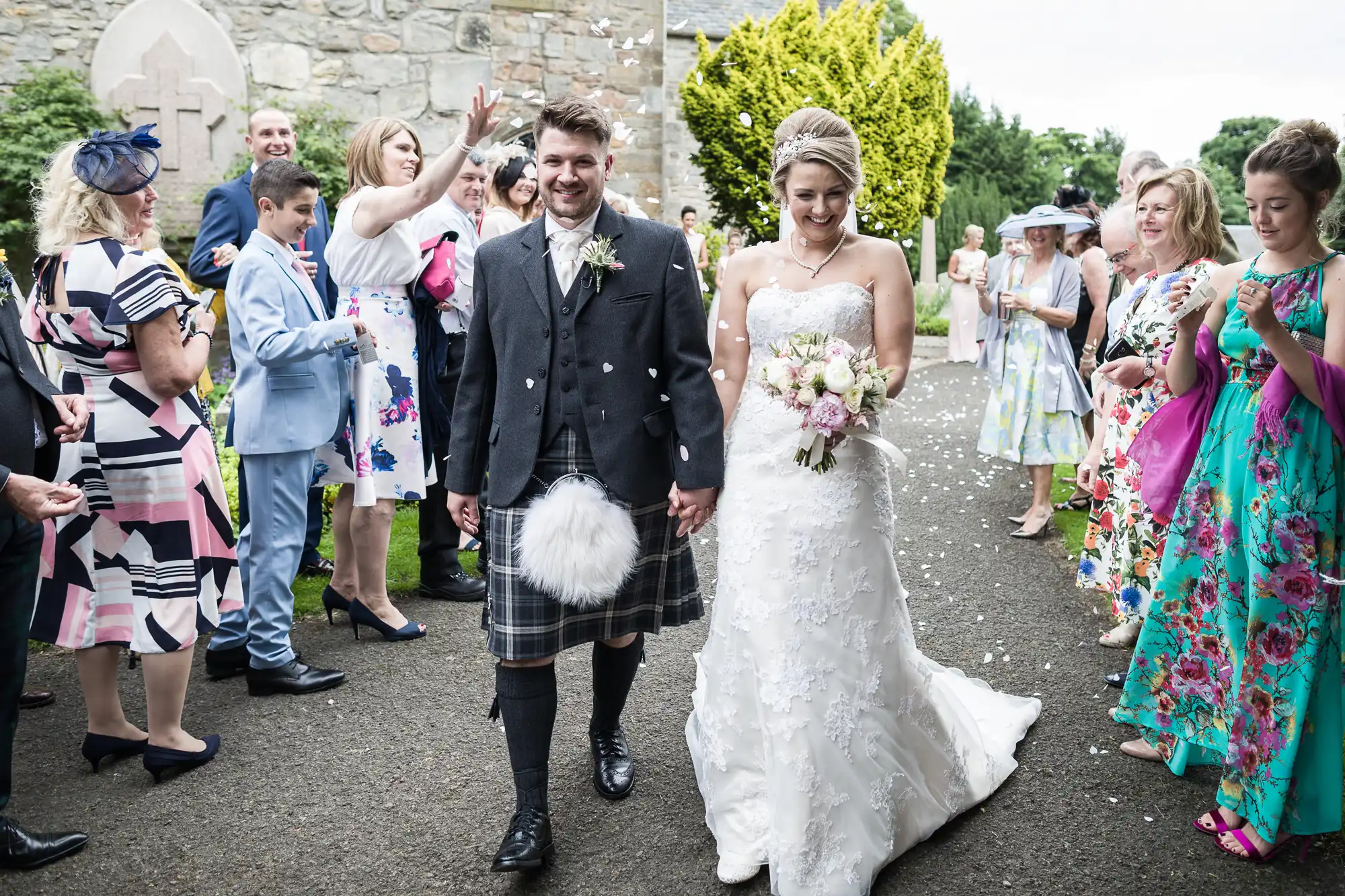 A groom in a kilt and a bride in a white dress walk hand-in-hand, surrounded by smiling guests outside a stone building.