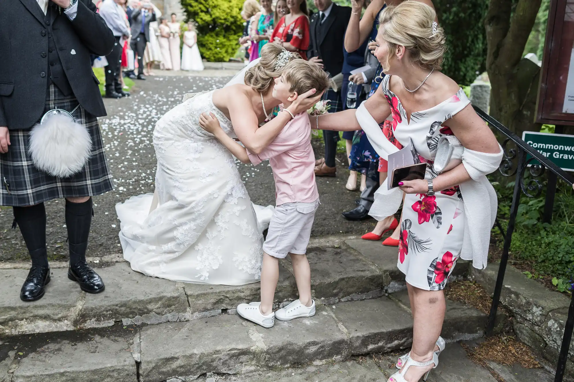 A bride in a white dress bending down to kiss a young boy while a woman in a floral dress stands beside them, holding a small box. Guests are visible in the background.