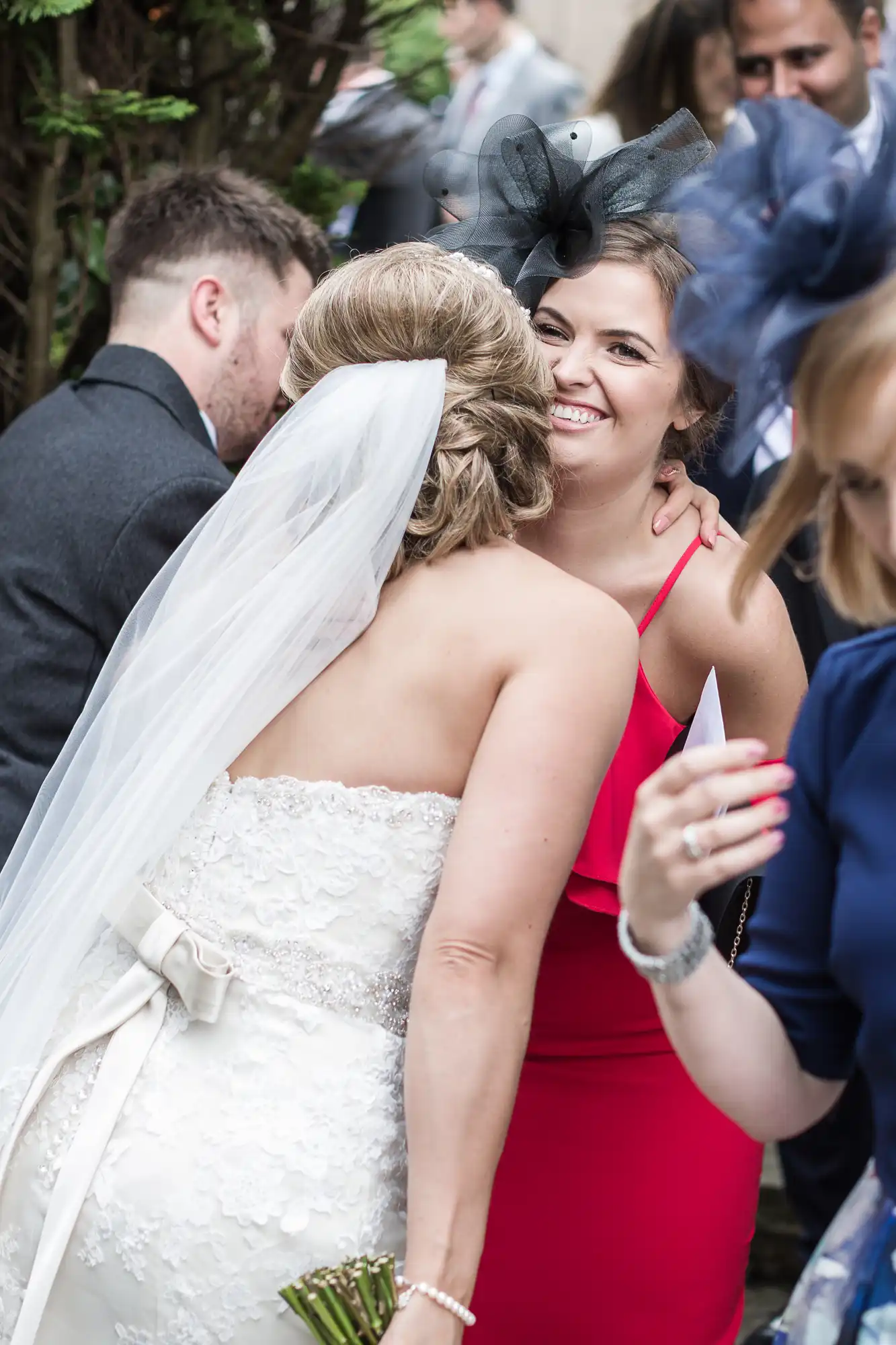 A bride in a strapless lace wedding dress with a long veil embraces a woman in a red dress at an outdoor event. Both are smiling, and other guests are visible in the background.