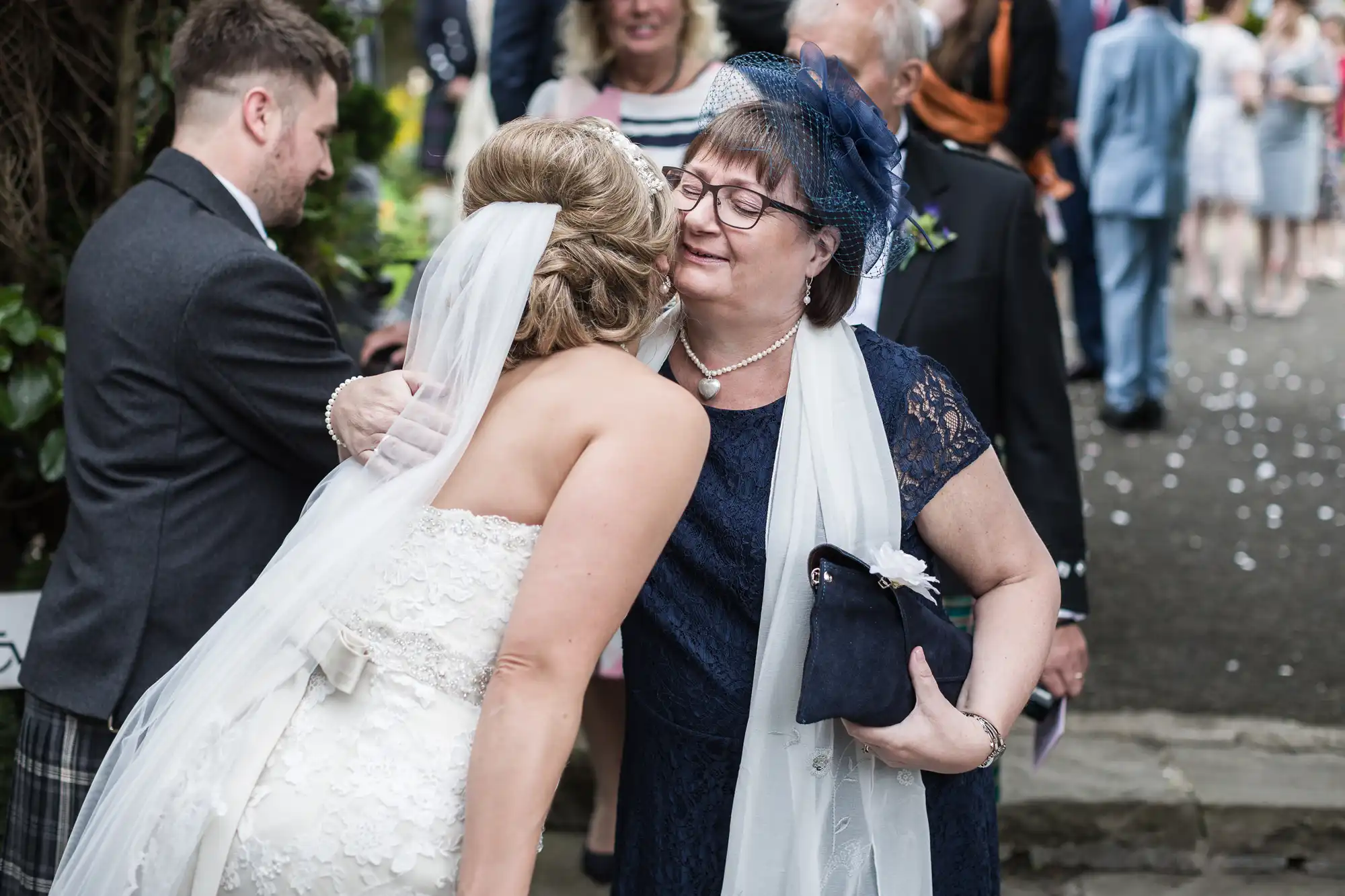 A bride in a white dress and veil hugs an older woman dressed in a navy blue lace outfit with matching hat during an outdoor wedding ceremony. Other guests are seen in the background.