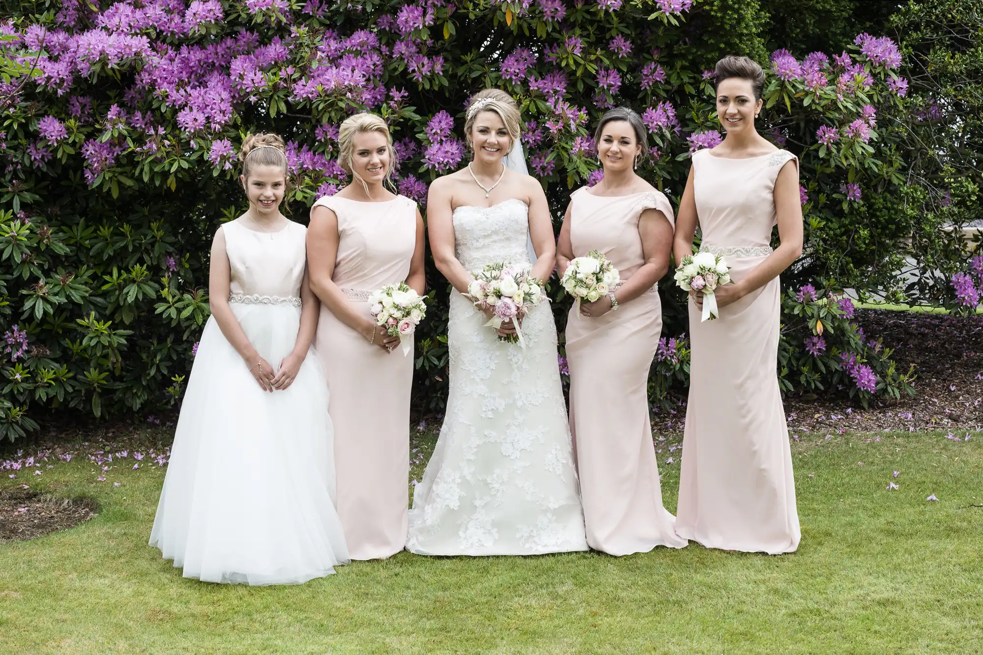 A bride in a white gown stands with four bridesmaids in light pink dresses, all holding bouquets, against a backdrop of blooming purple flowers.