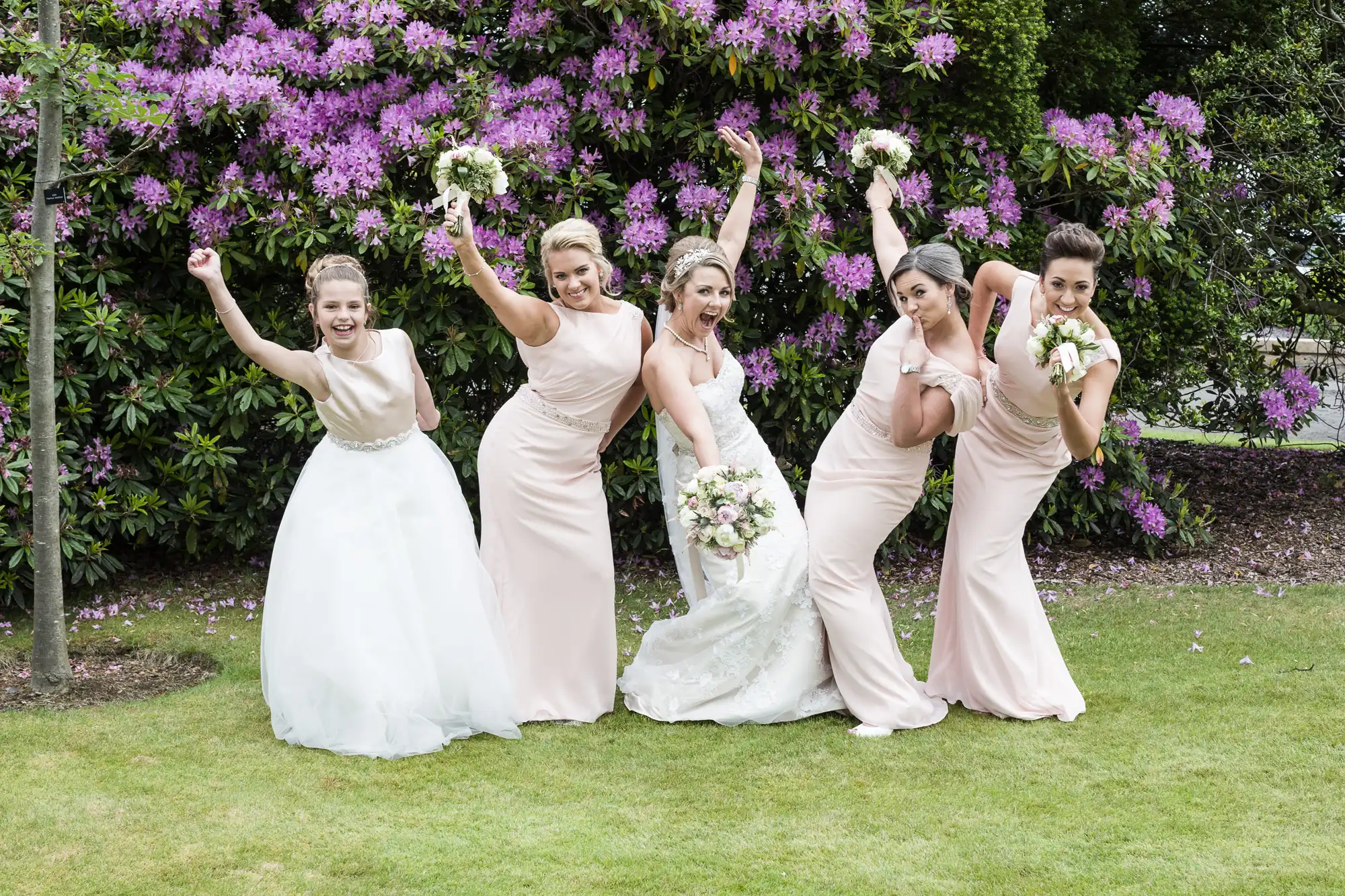 A bride and four bridesmaids in light pink dresses pose playfully in front of blooming purple flowers on a lawn.