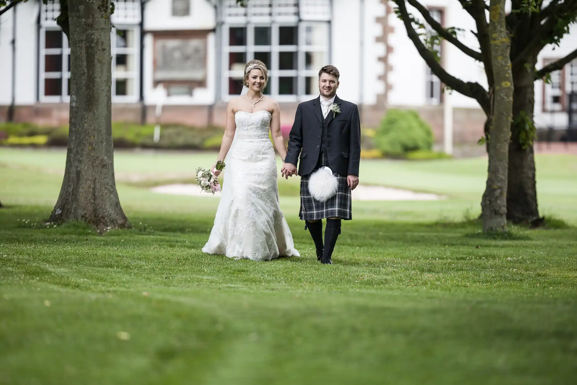 A bride in a white gown and a groom in a dark jacket with a kilt walk hand in hand through a grassy area with trees and a building in the background.