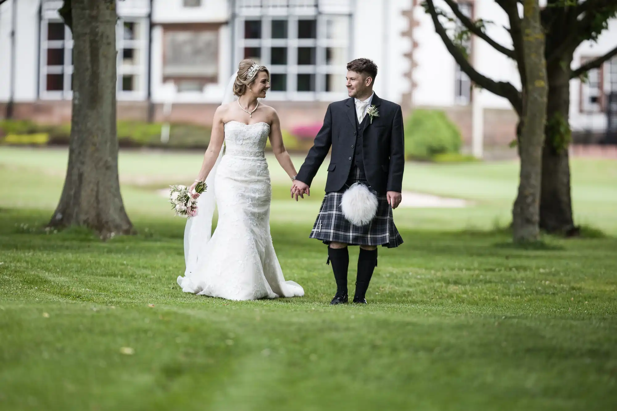 A bride in a white dress and a groom in a kilt walk hand-in-hand on a lawn with trees and a building in the background.