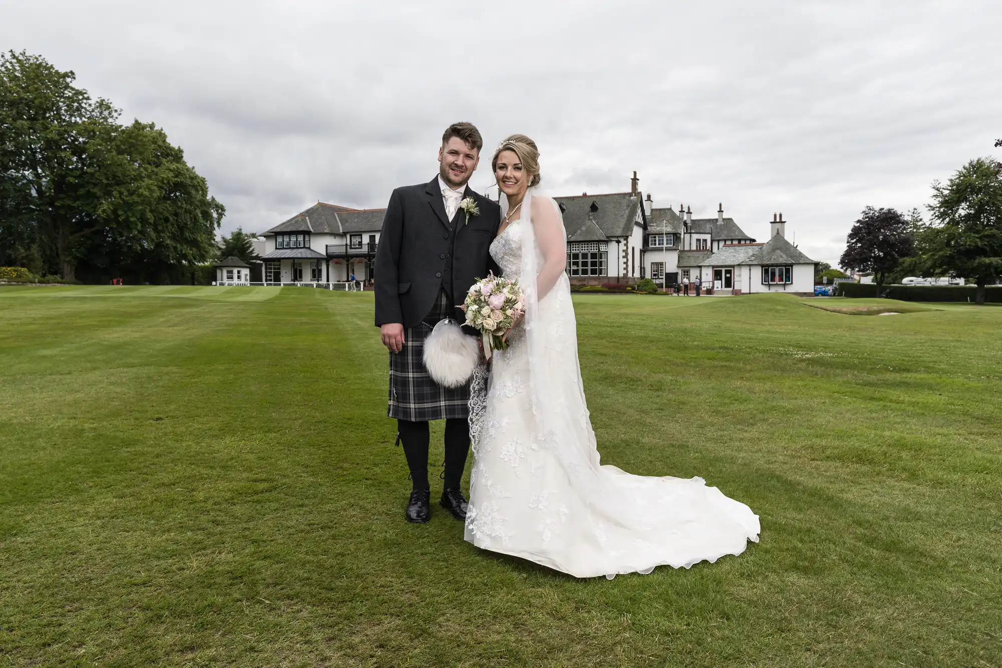 A bride in a white gown and a groom in a dark suit with a kilt stand on a lawn in front of a large, traditional building on a cloudy day.