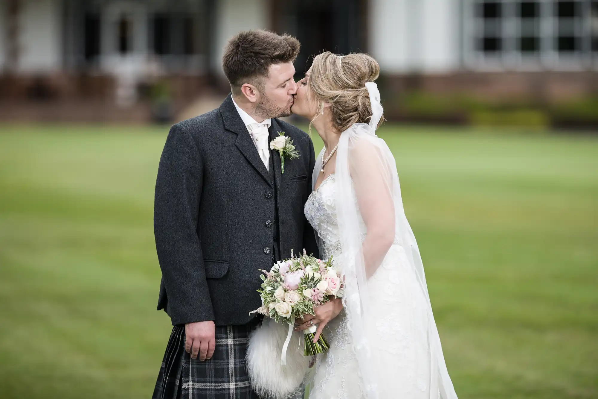 A bride and groom share a kiss on their wedding day. The groom is wearing a traditional Scottish kilt and jacket, while the bride is in a white dress and holding a bouquet.