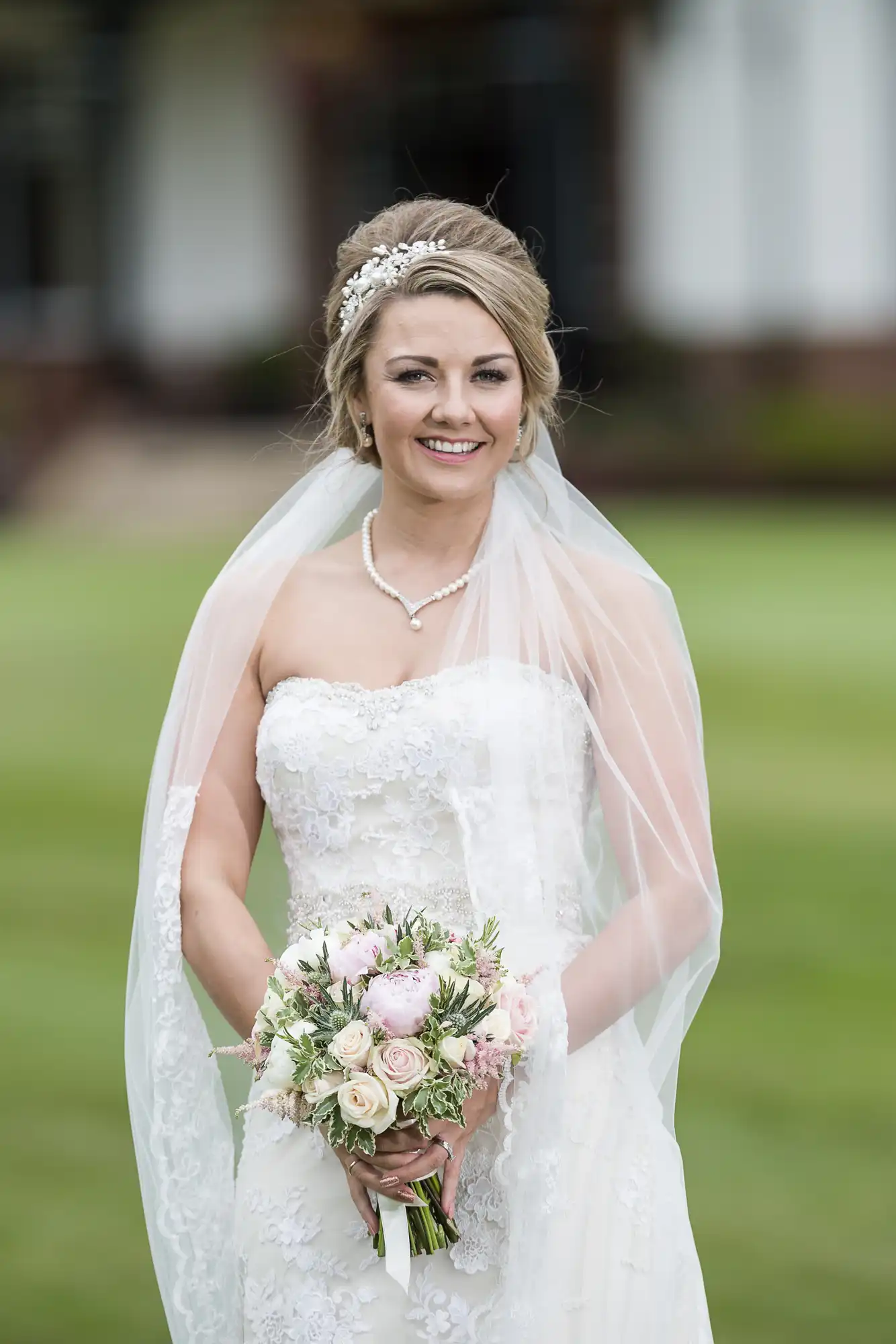 A bride in a strapless white wedding dress with a lace overlay and veil poses outdoors, holding a bouquet of flowers.