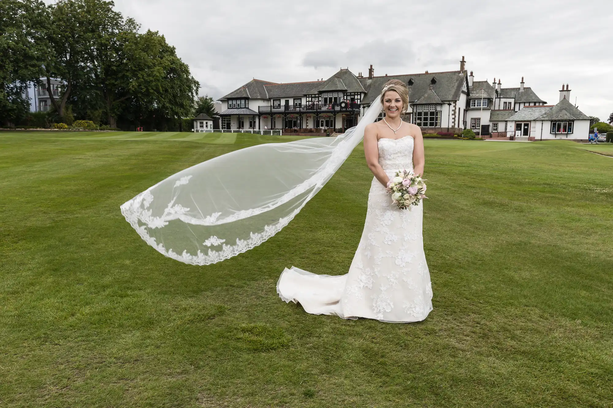 A bride in a white lace wedding dress and long veil stands on a grassy lawn in front of a large traditional building, holding a bouquet of flowers.