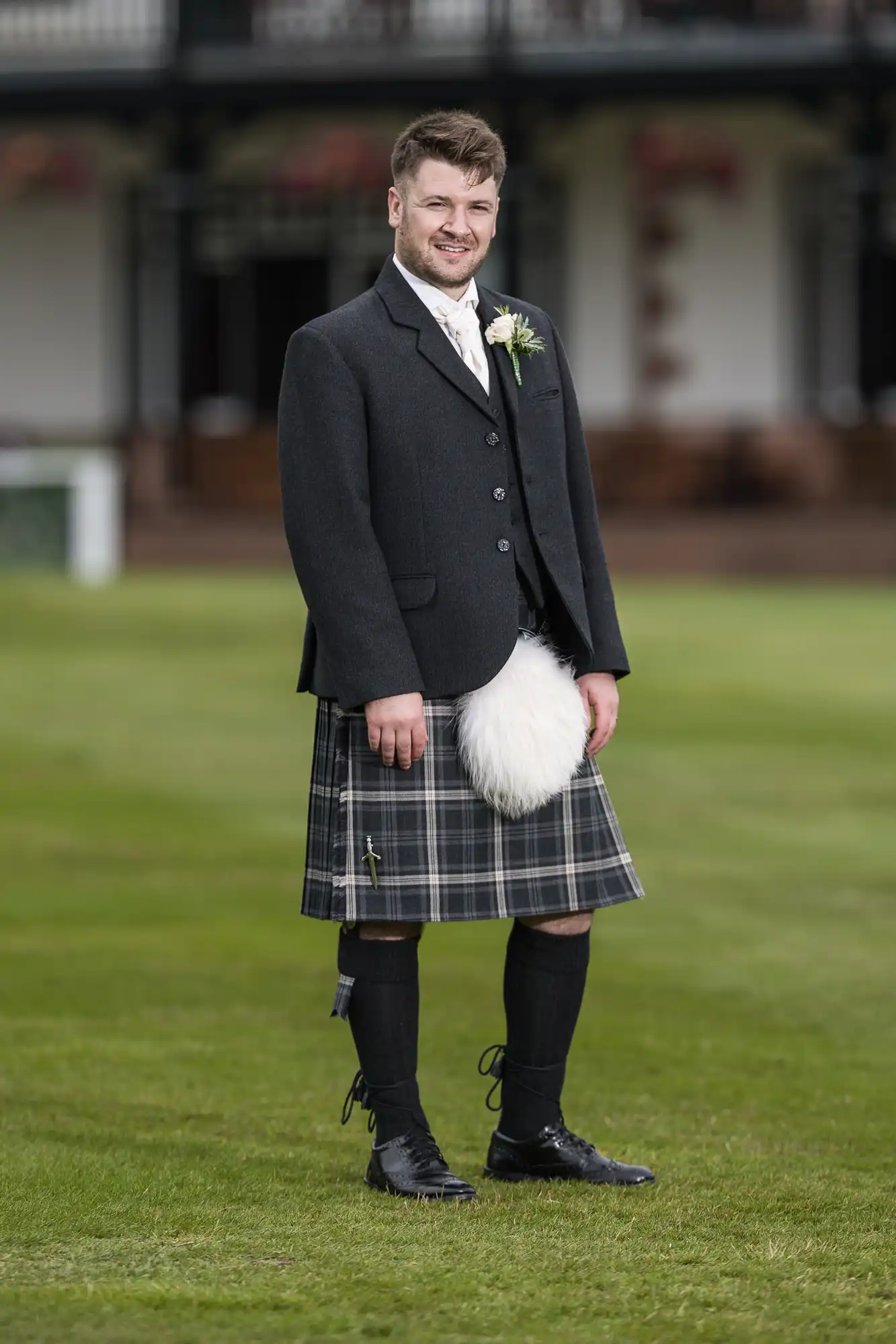 A man stands on grass wearing a black jacket, white shirt, and plaid kilt, with a sporran and boutonniere, and knee-high socks with shoes.