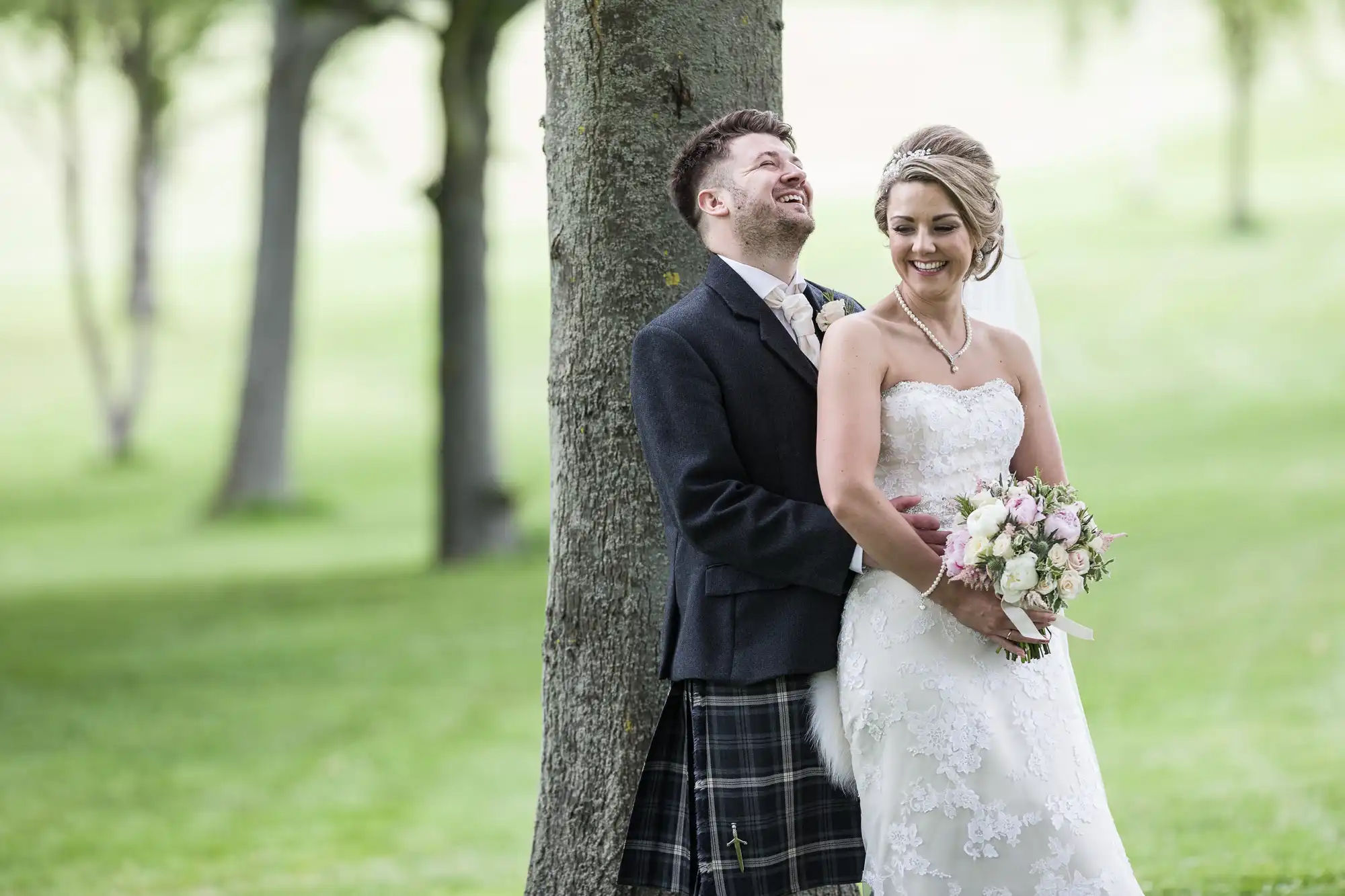 A couple dressed in wedding attire stands outdoors. The groom is wearing a kilt and jacket, while the bride is in a strapless white dress holding a bouquet. Both are smiling and standing next to a tree.