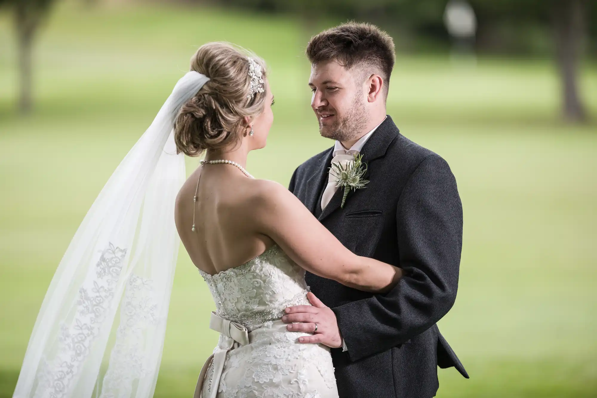 A bride and groom stand together outdoors, dressed in wedding attire. The bride's back is to the camera as she holds the groom, who is smiling and looking at her.