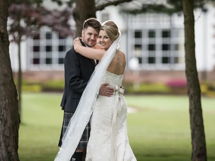 Cramond Kirk wedding: newlyweds embrace under trees, with the bride wearing a white gown and veil, and the groom in a dark suit, in front of a building with large windows.